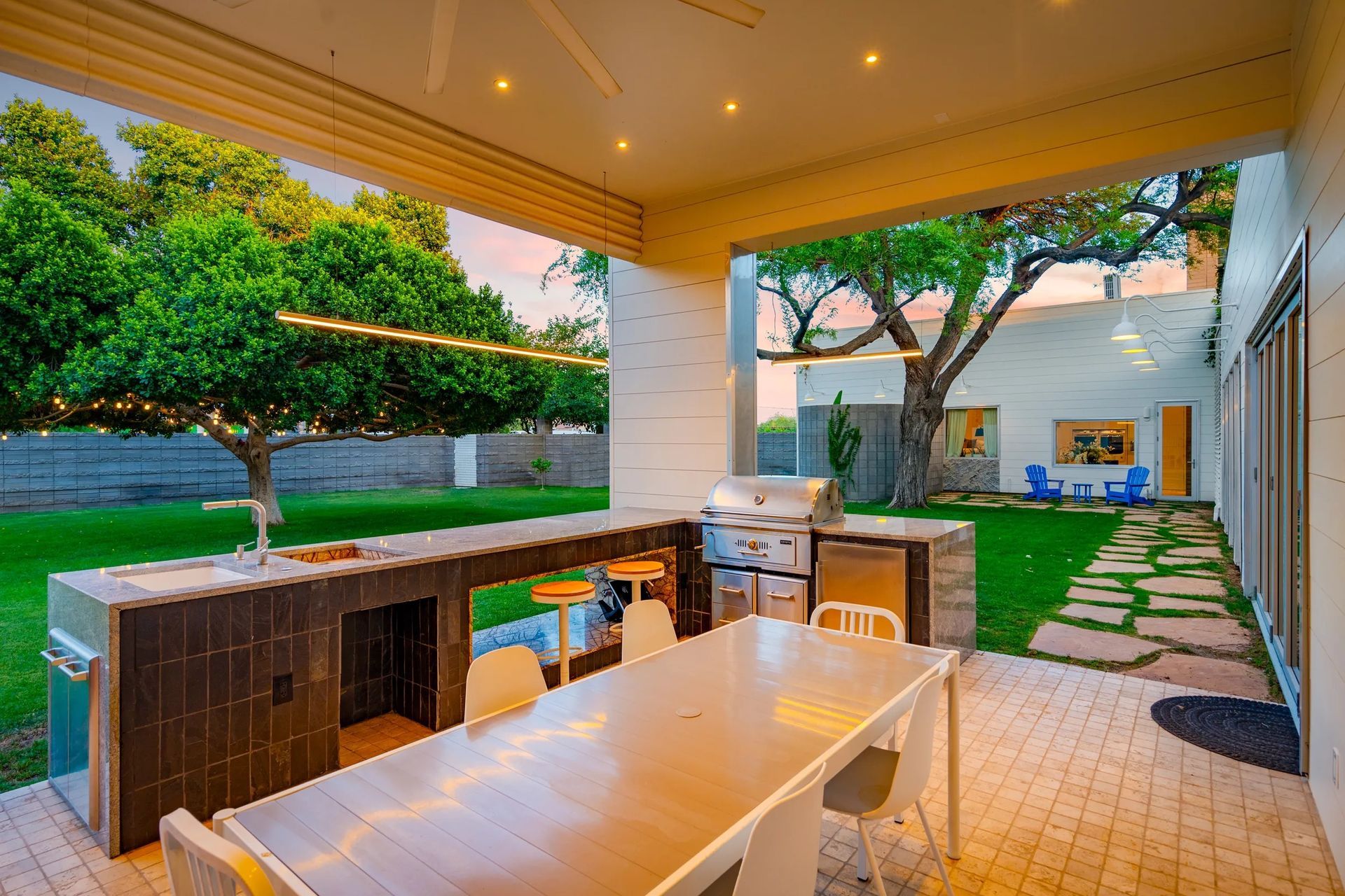 Covered outdoor kitchen with a grill, bar, and dining table; green lawn and trees in the background.