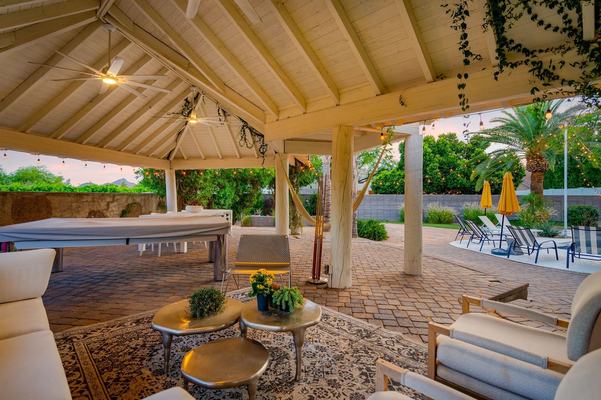 A luxurious outdoor patio with a white pergola, seating areas, and a pool table. Greenery surrounds the space, and the color palette is neutral.