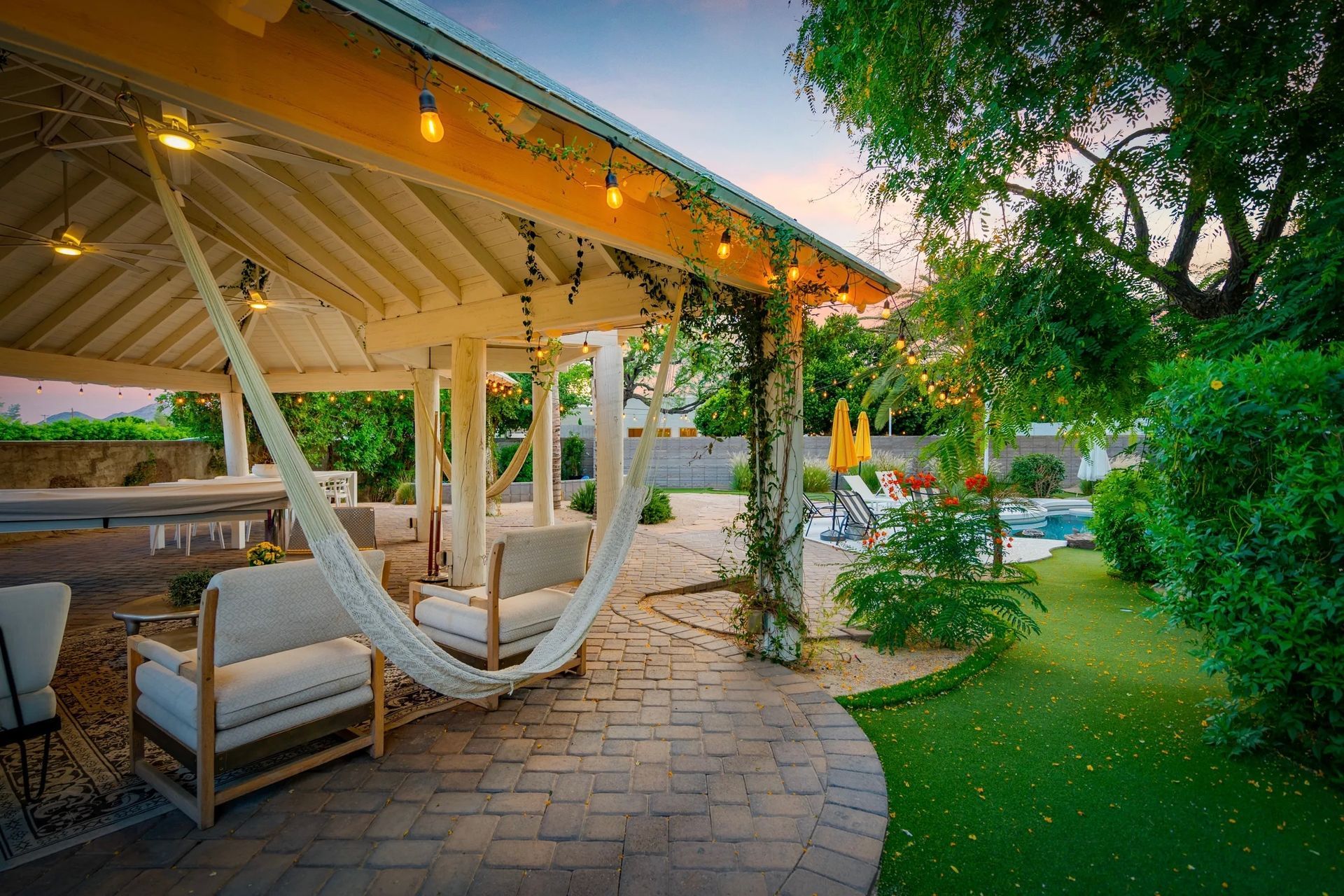 A patio with a hammock, seating, and string lights, overlooking a lawn and pool. Trees surround the area at sunset.