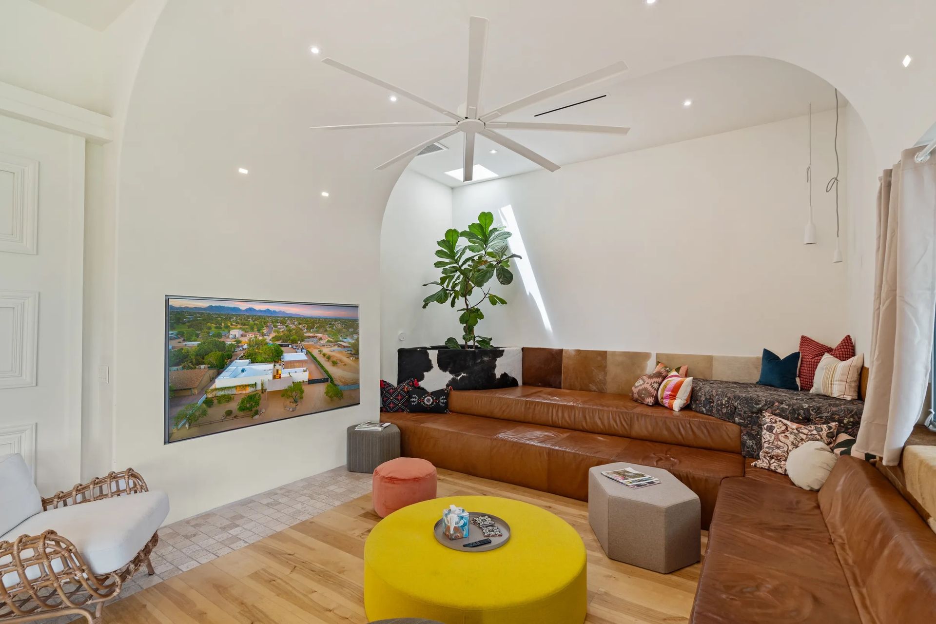 Bright living room with tiered leather seating, round yellow ottoman, large screen TV, and oversized ceiling fan.