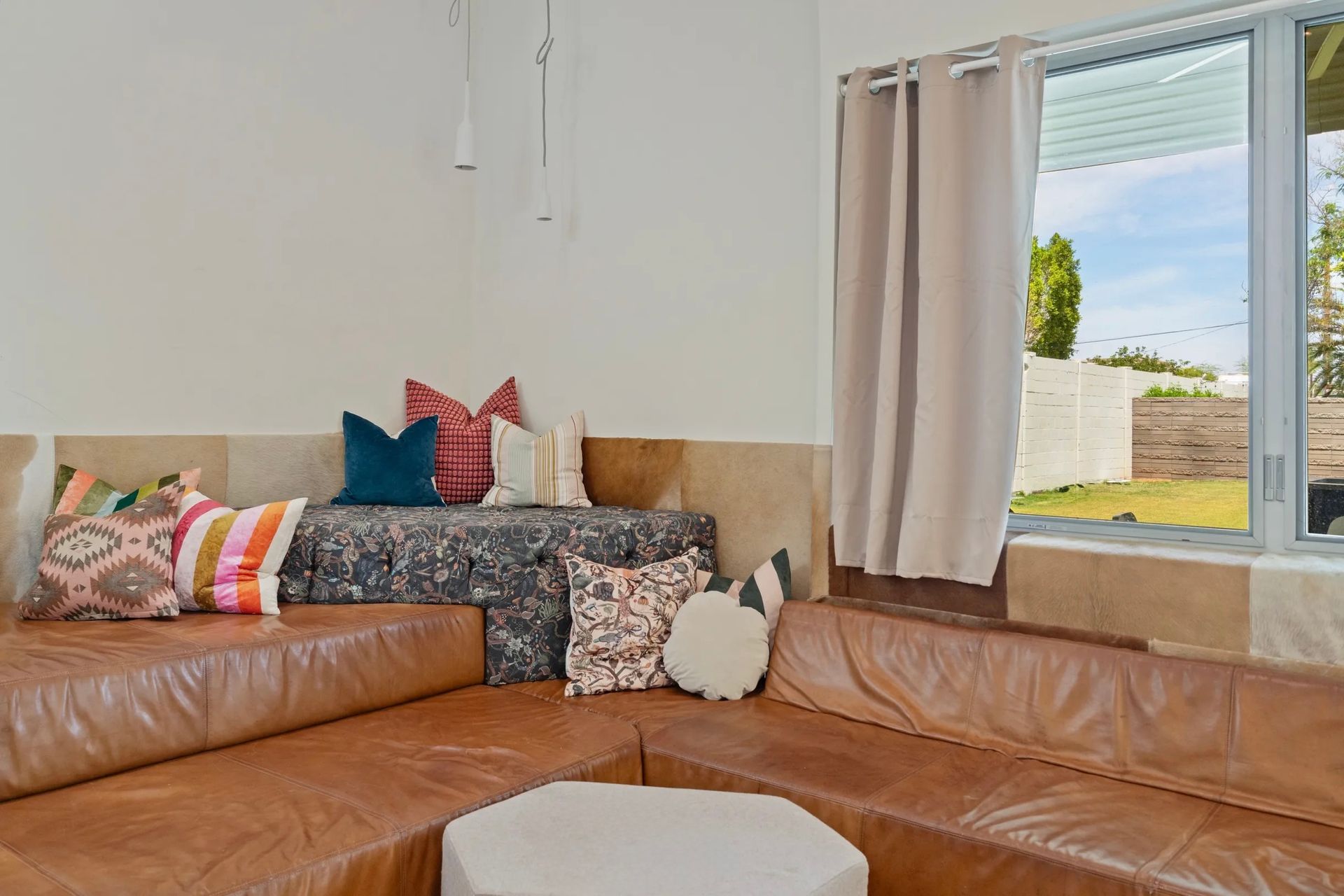 A cozy, sunlit living space with a brown leather sectional couch and colorful throw pillows. A window with curtains overlooks a yard.