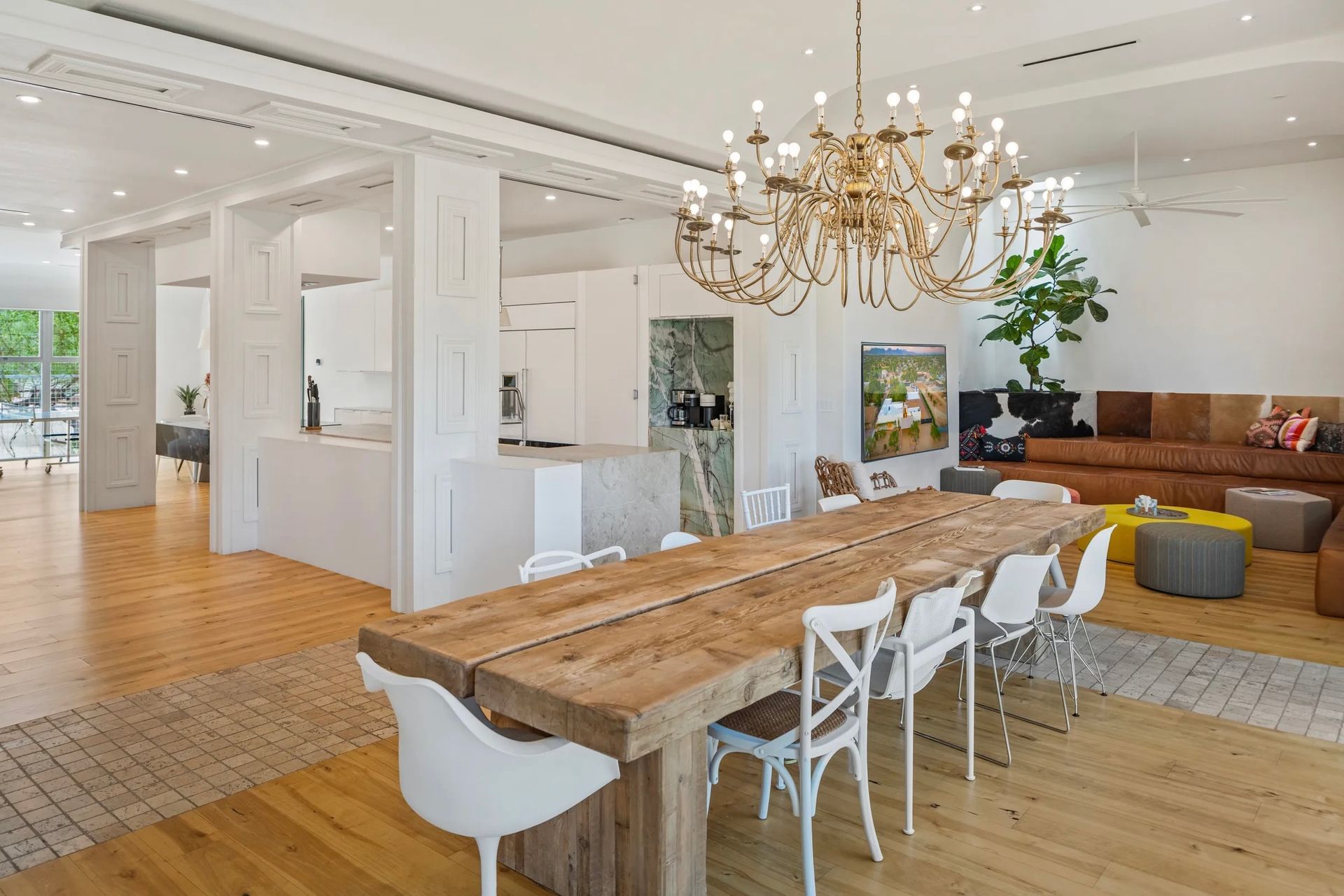 Spacious, light-filled dining and living area. Features a long wooden dining table, a large chandelier, and a brown leather sofa.