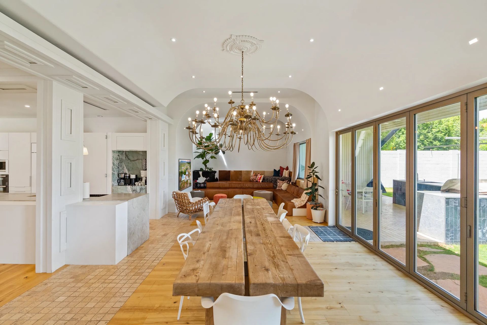 A long, wooden dining table in a bright, open room with a large chandelier and glass doors leading to a yard.