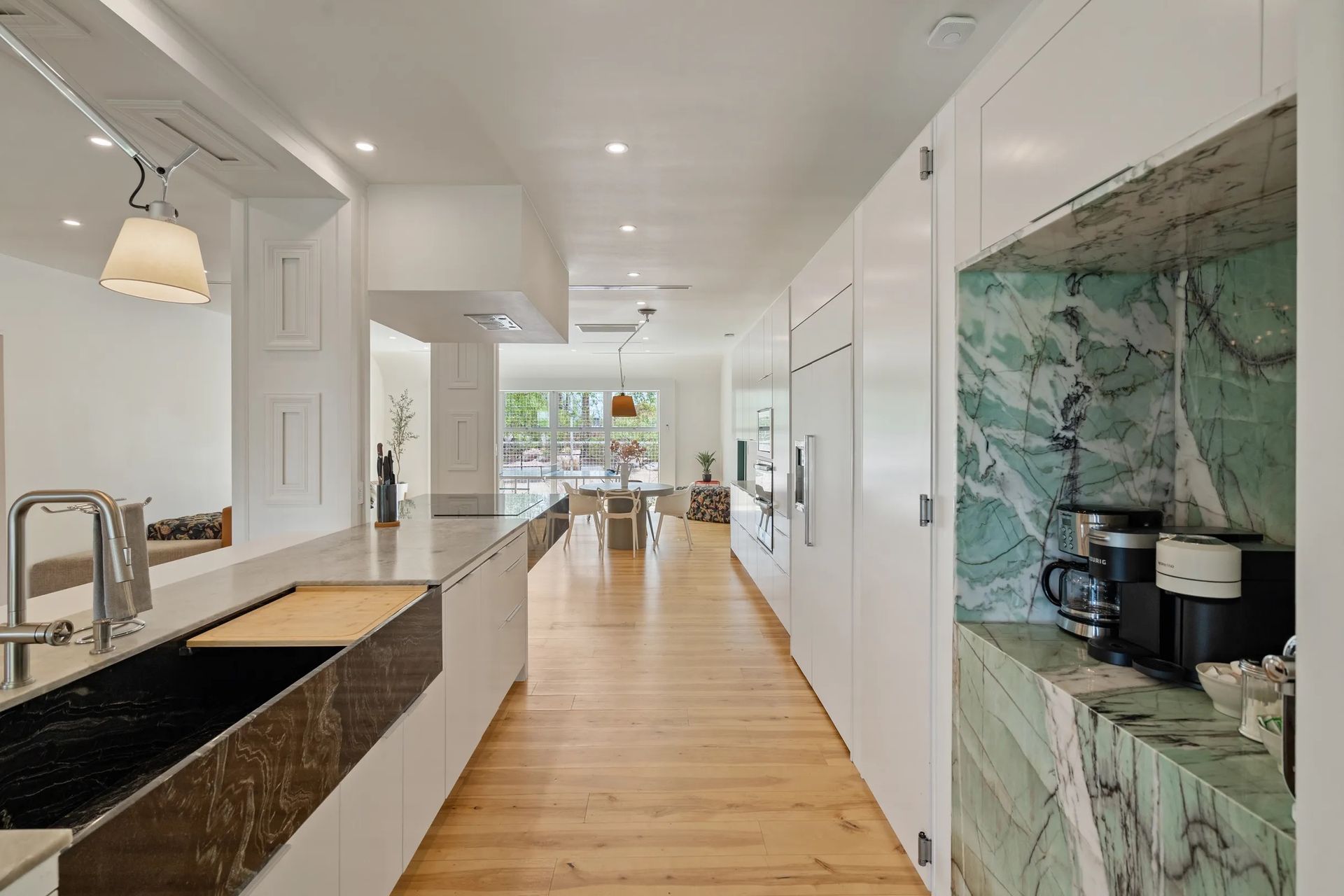 Long, modern kitchen with light wood floors, white cabinets, and a green marble accent. A long island and a dining area are visible in the background.