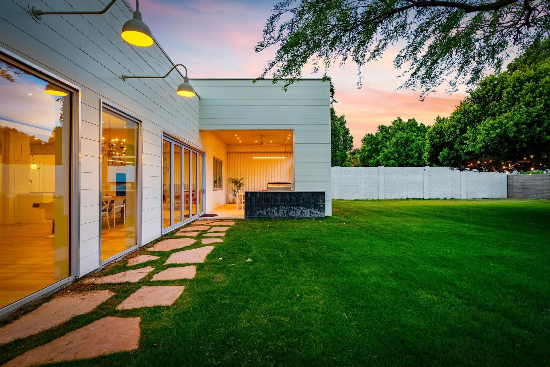 Modern home exterior with a green lawn, stone pathway, large windows, and yellow exterior lights at sunset.