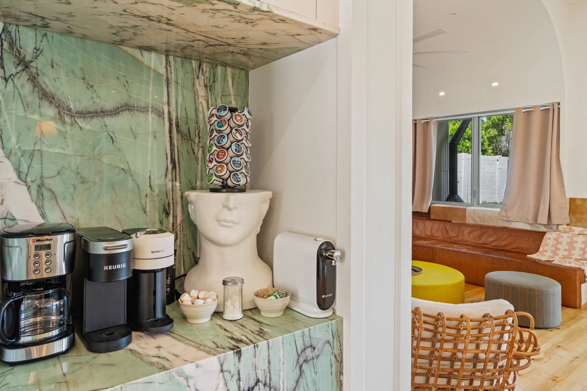 Coffee station with green marble countertop, coffee machines, decorative items, and a glimpse of a living area.