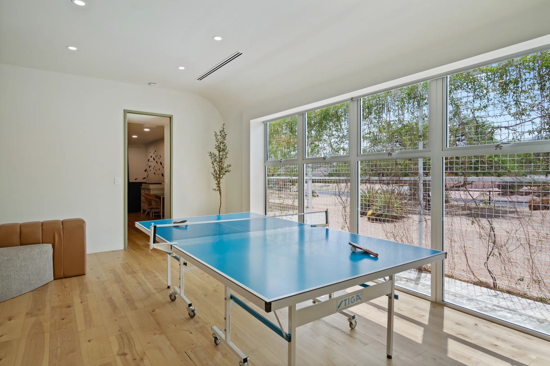A bright room with a blue ping pong table, large windows, and a doorway to another room. Sunlight streams in, and the floor is wood.