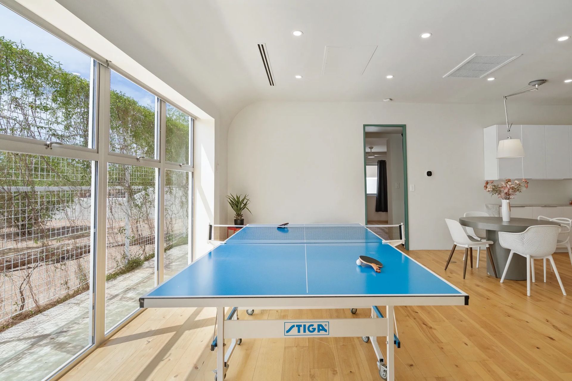 A bright room with a blue ping pong table, large windows, and a dining area with white furniture.
