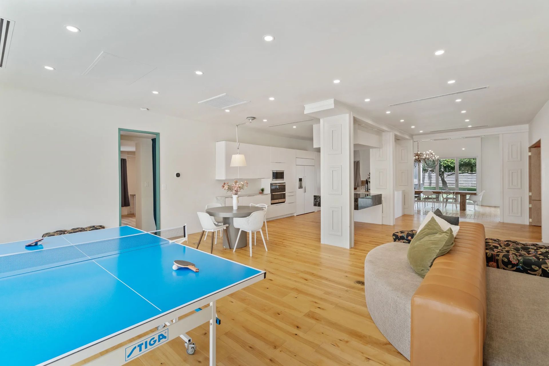 Spacious modern apartment interior with ping pong table, kitchen, living area, and outdoor access. Bright, white walls and wood floors.