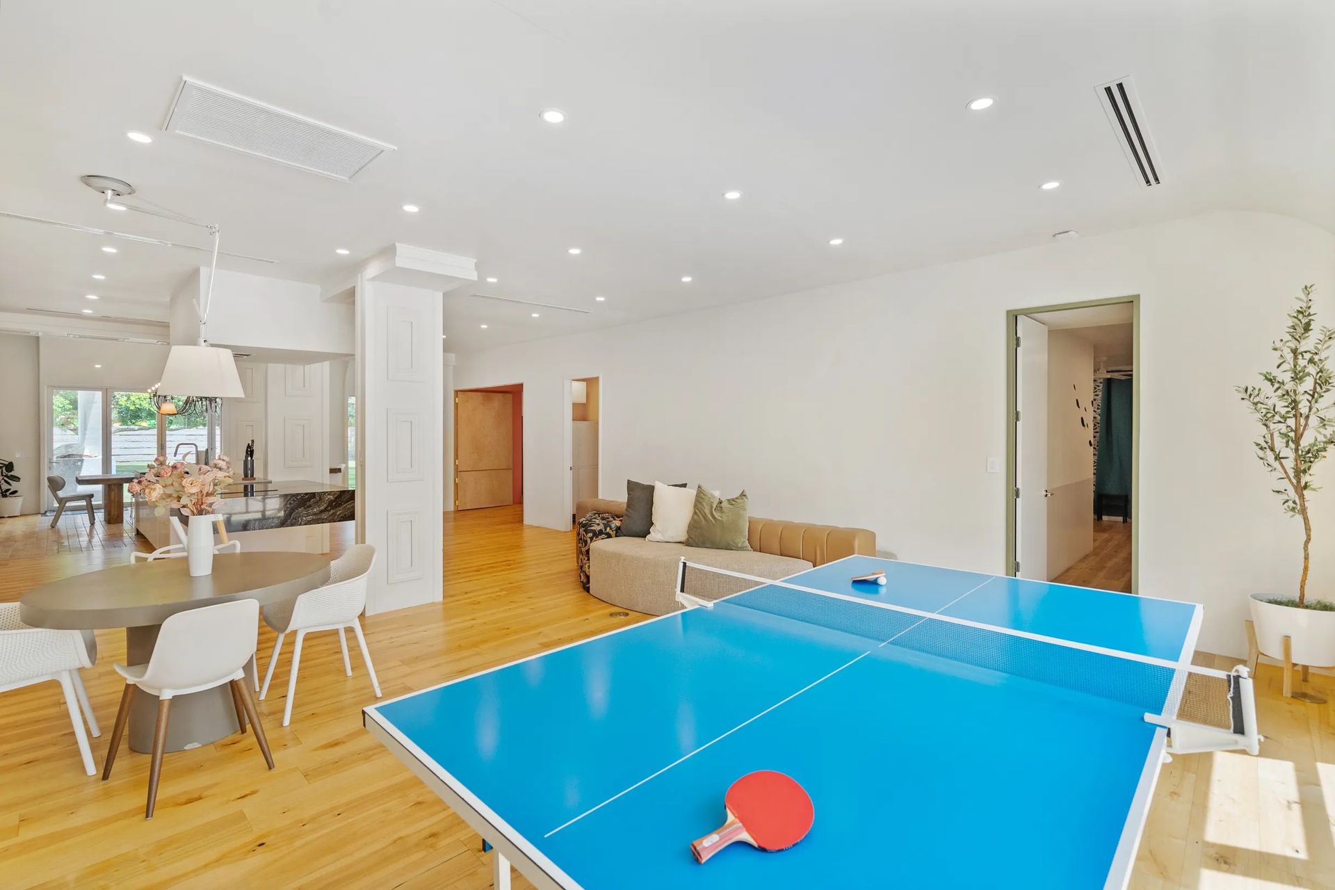 Spacious modern room with a blue ping pong table, dining table, and a seating area. Light-colored wood floors and white walls create a bright atmosphere.