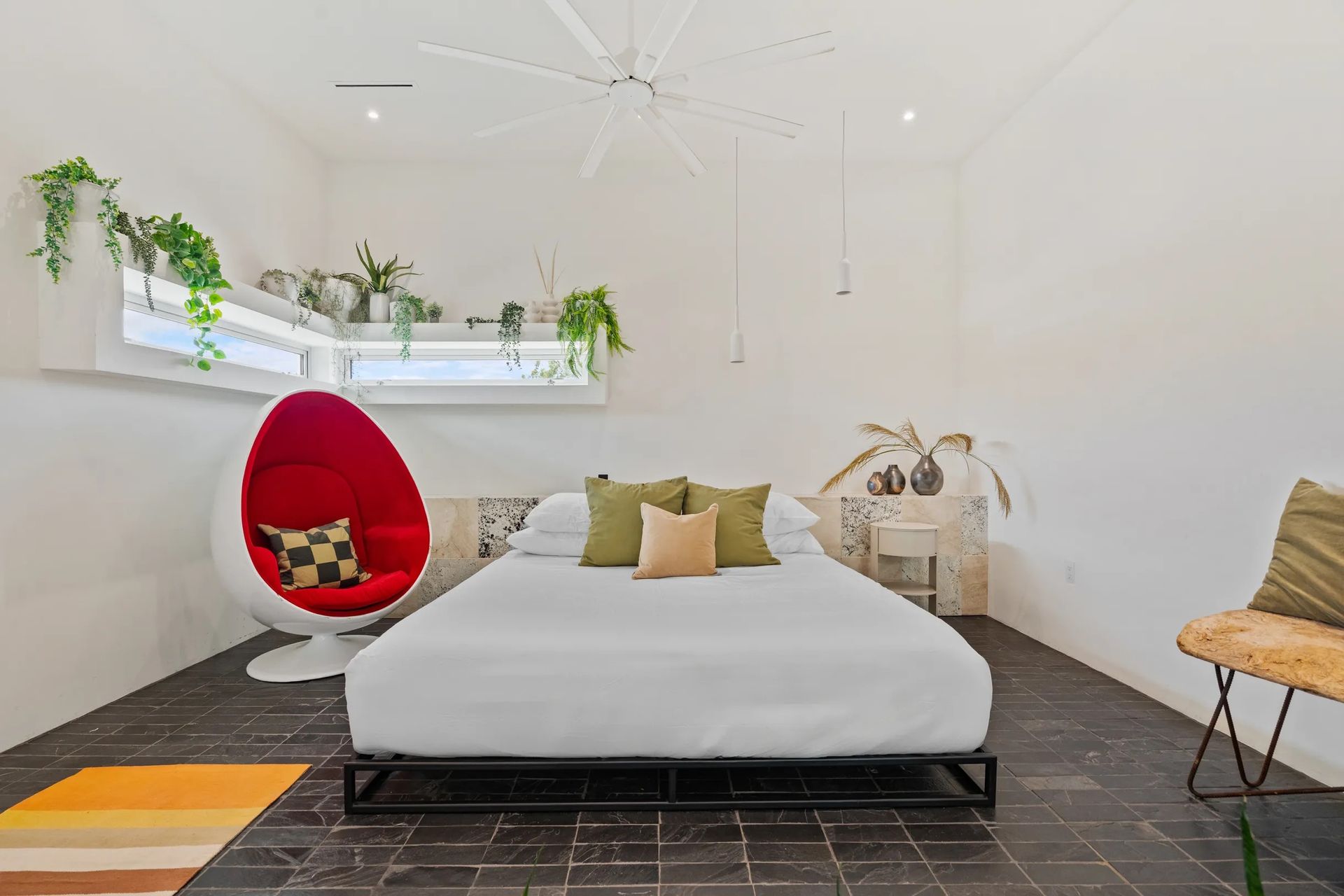 Modern bedroom with white walls, black floor, and a red egg chair. The room has a bed, plants, and a geometric rug.