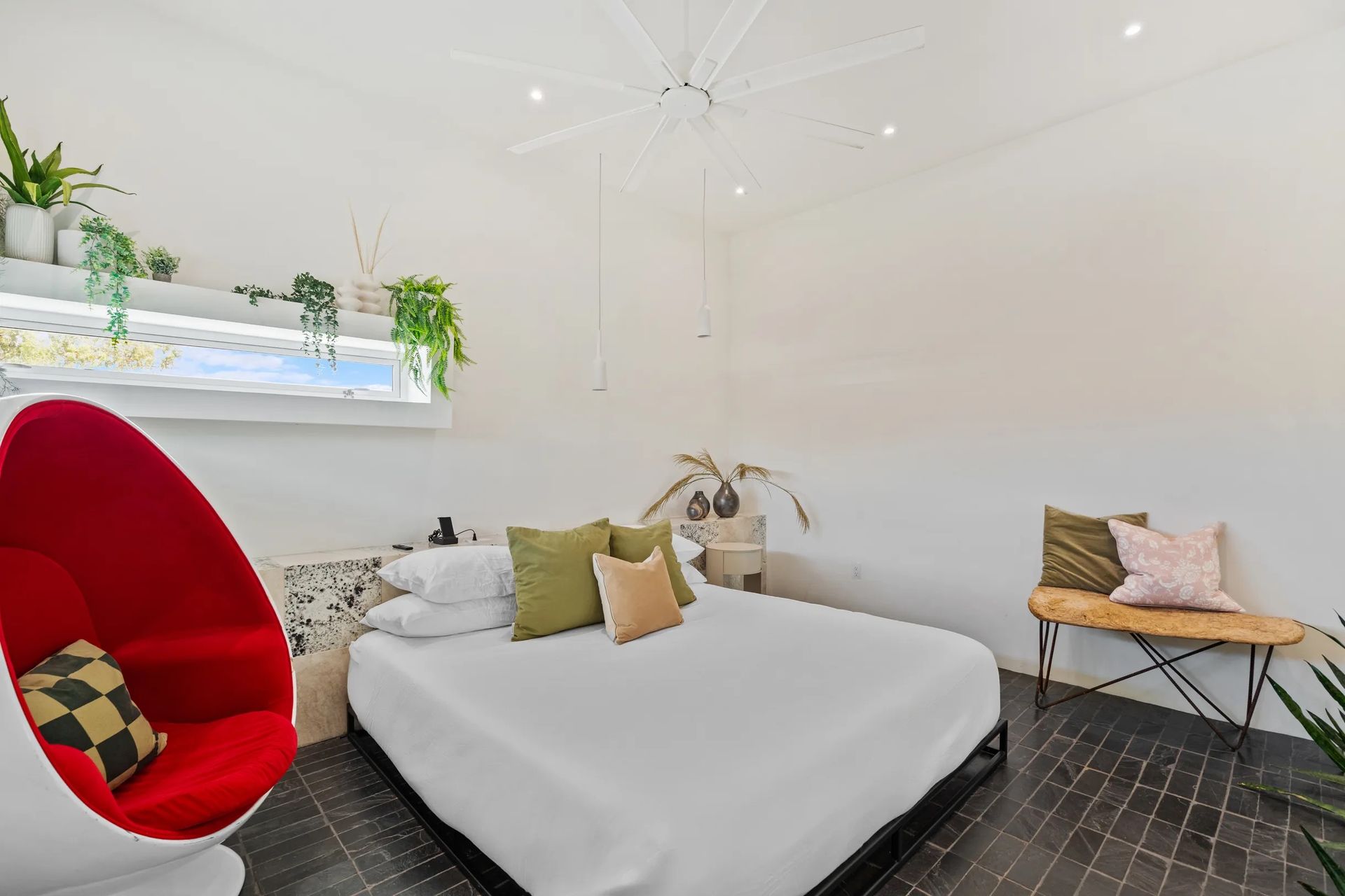 Bright bedroom with a red egg-shaped chair, bed with green pillows, and plants on a shelf.