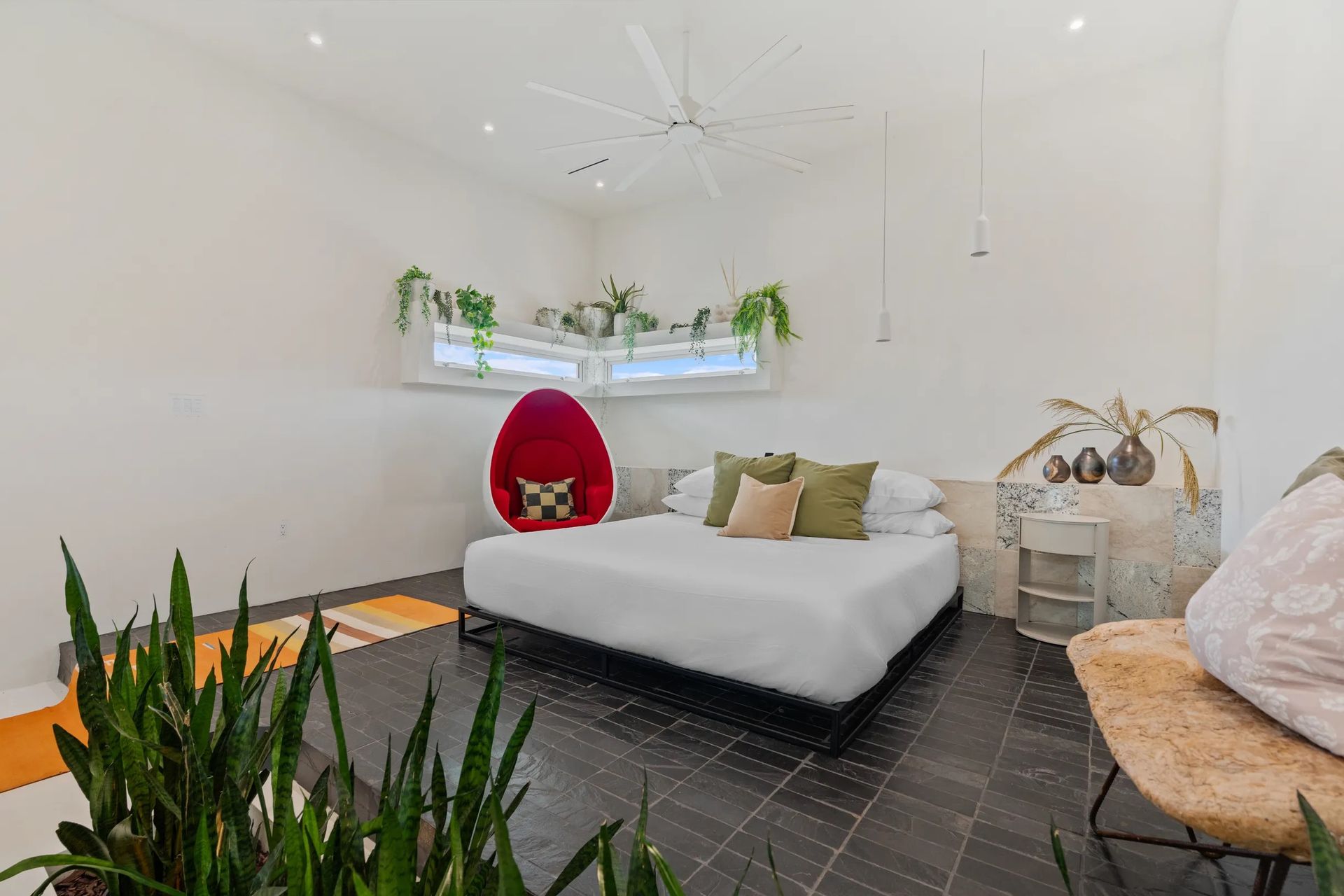 A bright bedroom with a bed, plants, and a large egg chair. White walls, dark floor, and a large window.