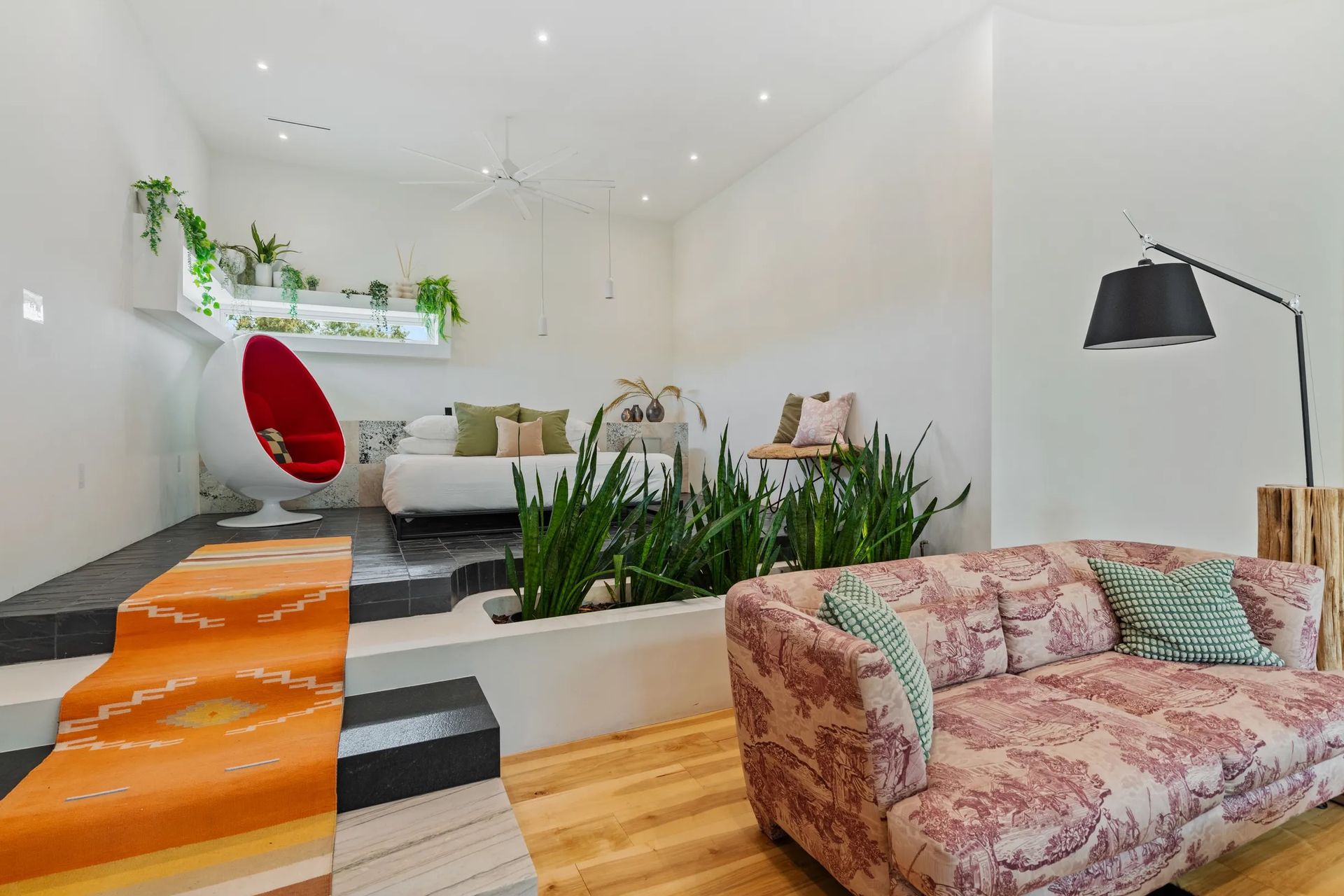 Cozy, modern living space with plants, seating areas, and a red hanging chair. Features white walls, a rug, and wood and black tile accents.