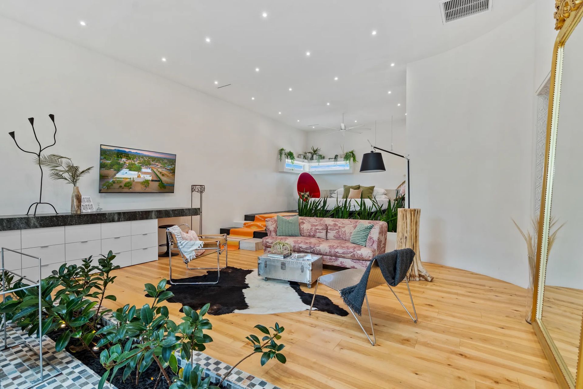 Bright, spacious room with hardwood floors, white walls, and plants. Features a floral sofa, gold mirror, and a black and white cowhide rug.