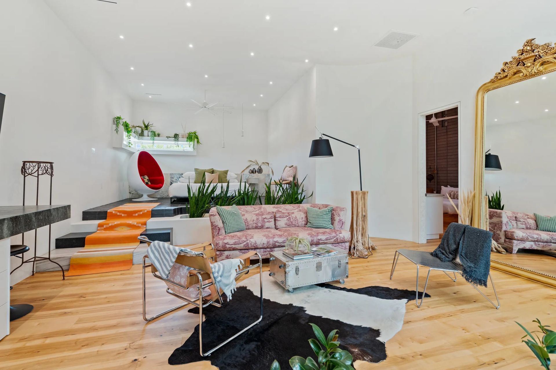 Spacious, brightly lit room with a pink floral sofa, various seating options, and a large gold-framed mirror.