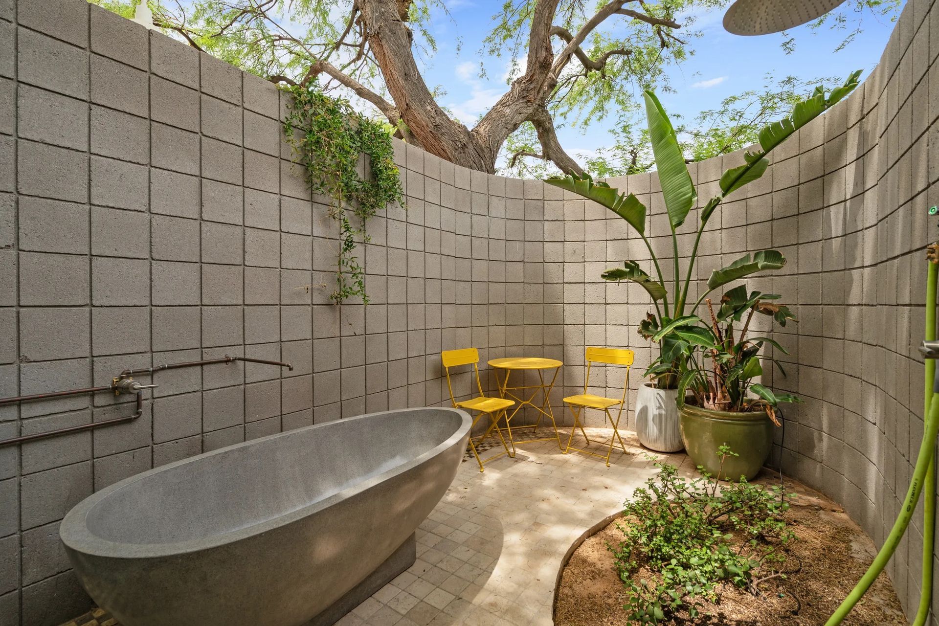 Outdoor shower area with gray concrete block walls, a stone tub, yellow chairs, and lush greenery.