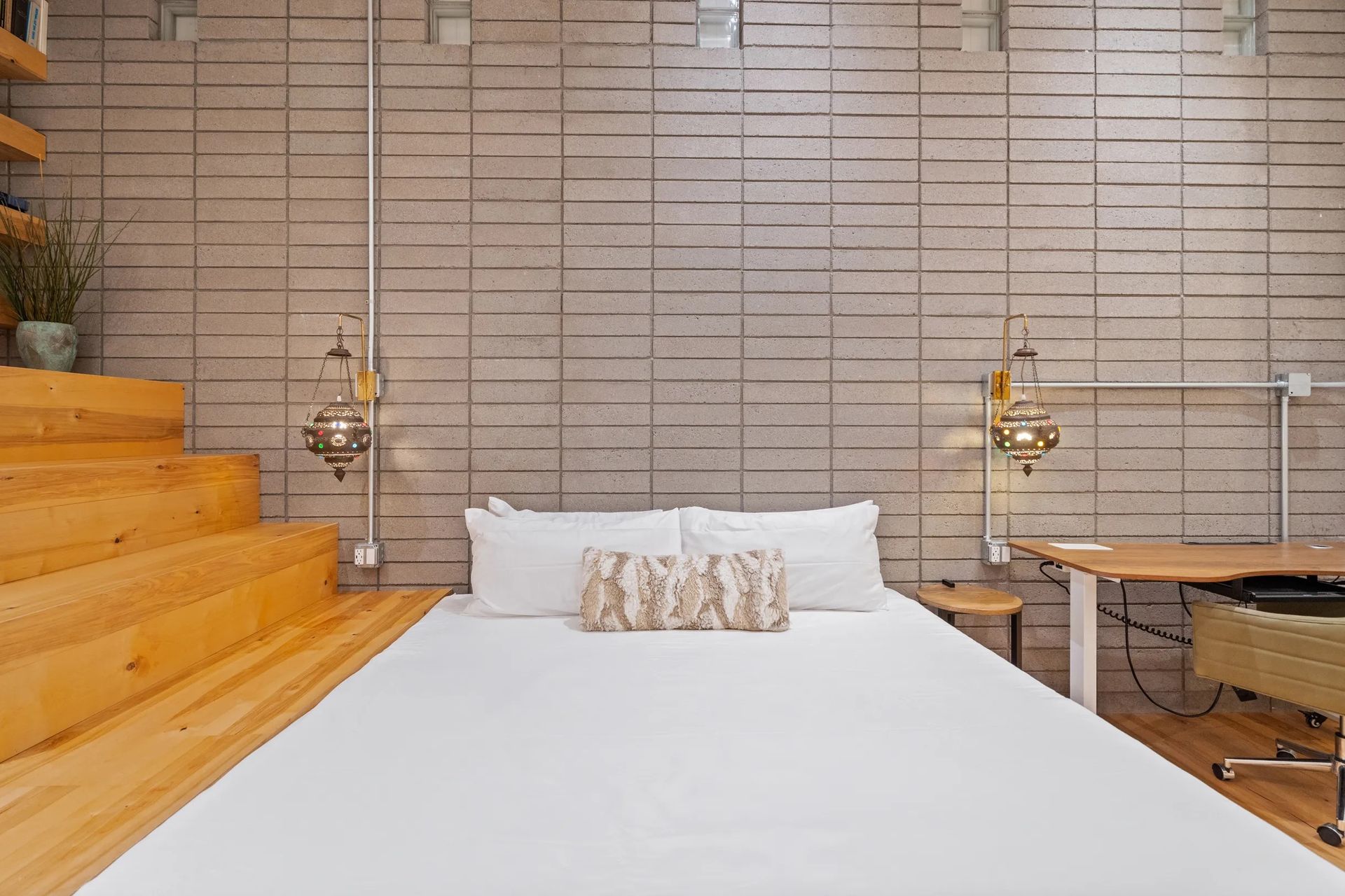 Bedroom with a white-sheeted bed, wooden stairs, and concrete block wall with metal light fixtures.