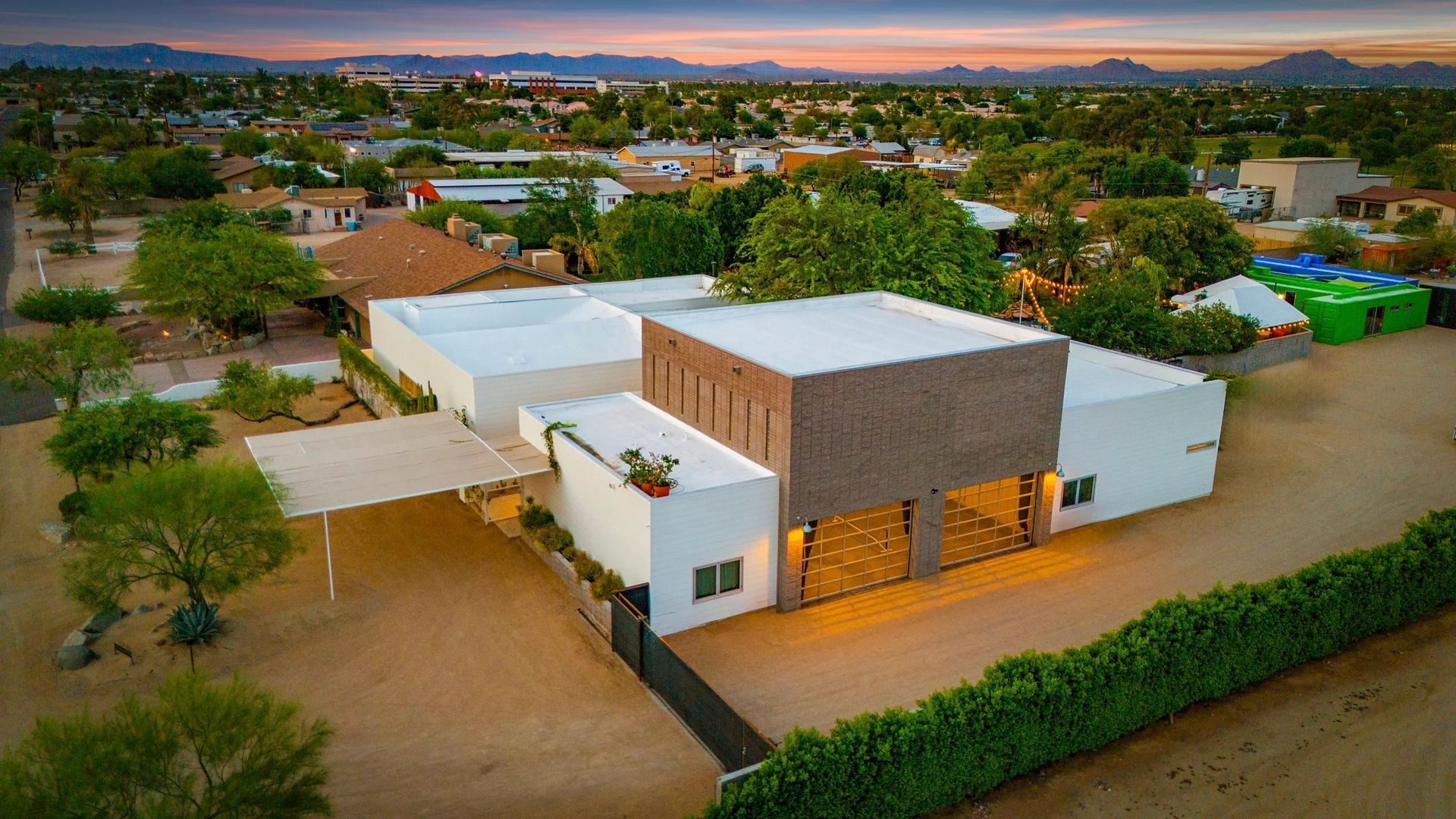 Aerial view of a modern, white house with a brown facade and a covered patio in a desert setting; sunset in the background.