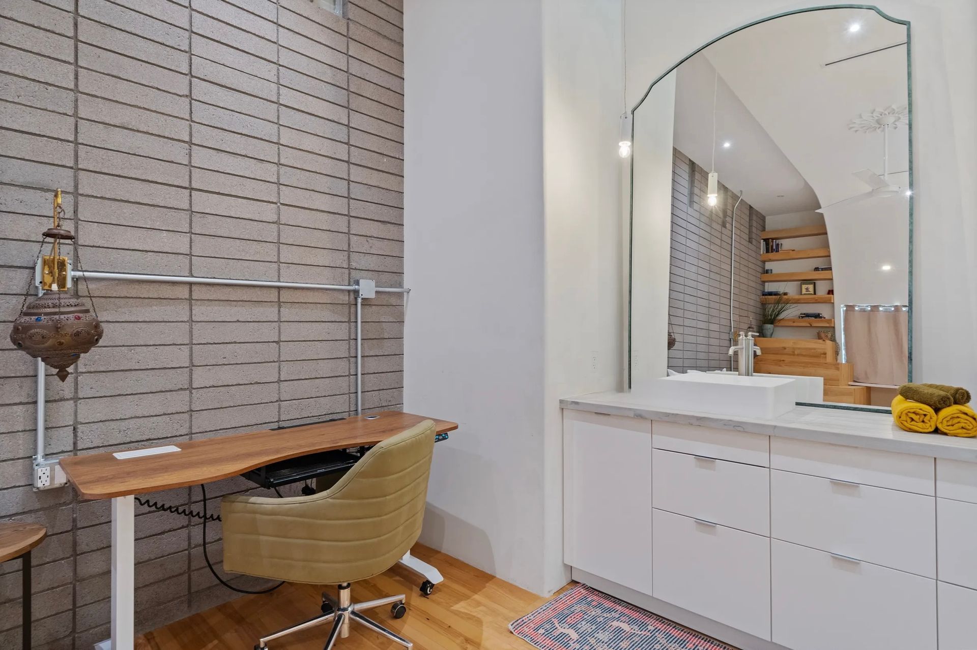 A vanity area with a desk, mirror, and sink. Neutral tones dominate, with a patterned accent rug on the wooden floor.