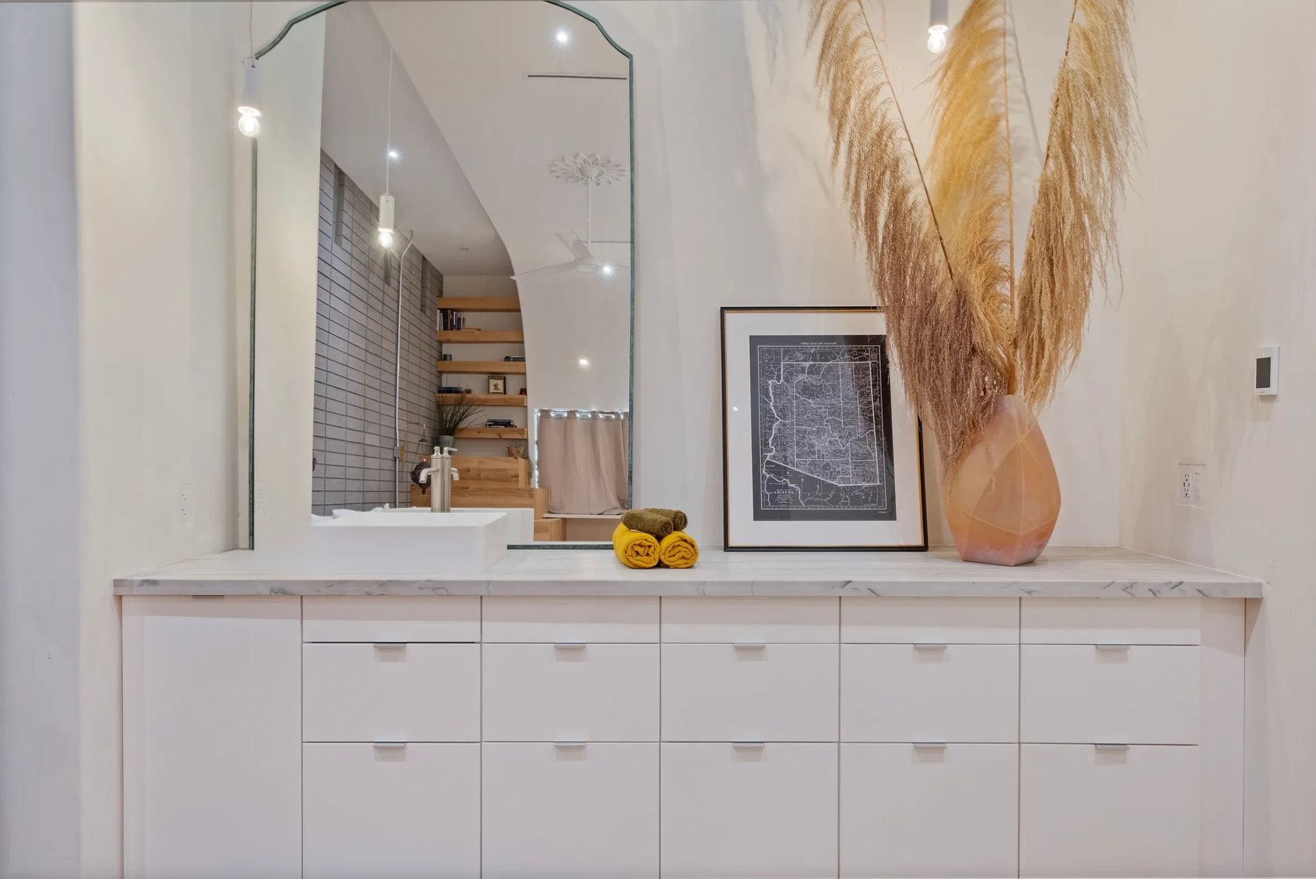 A modern entryway with a large arched mirror, white cabinets, and decorative vase with tall dried grasses.