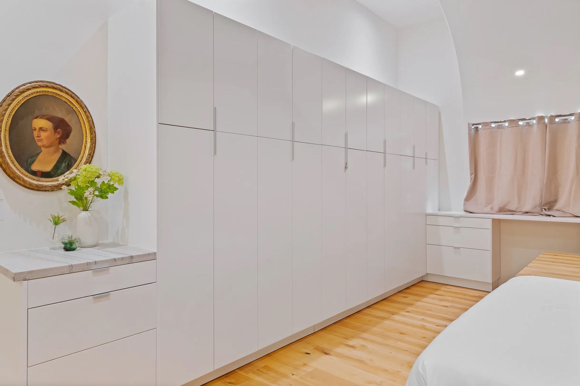 Bedroom with a large white closet, hardwood floors, and a painting on the wall. There's a window with curtains in the background.