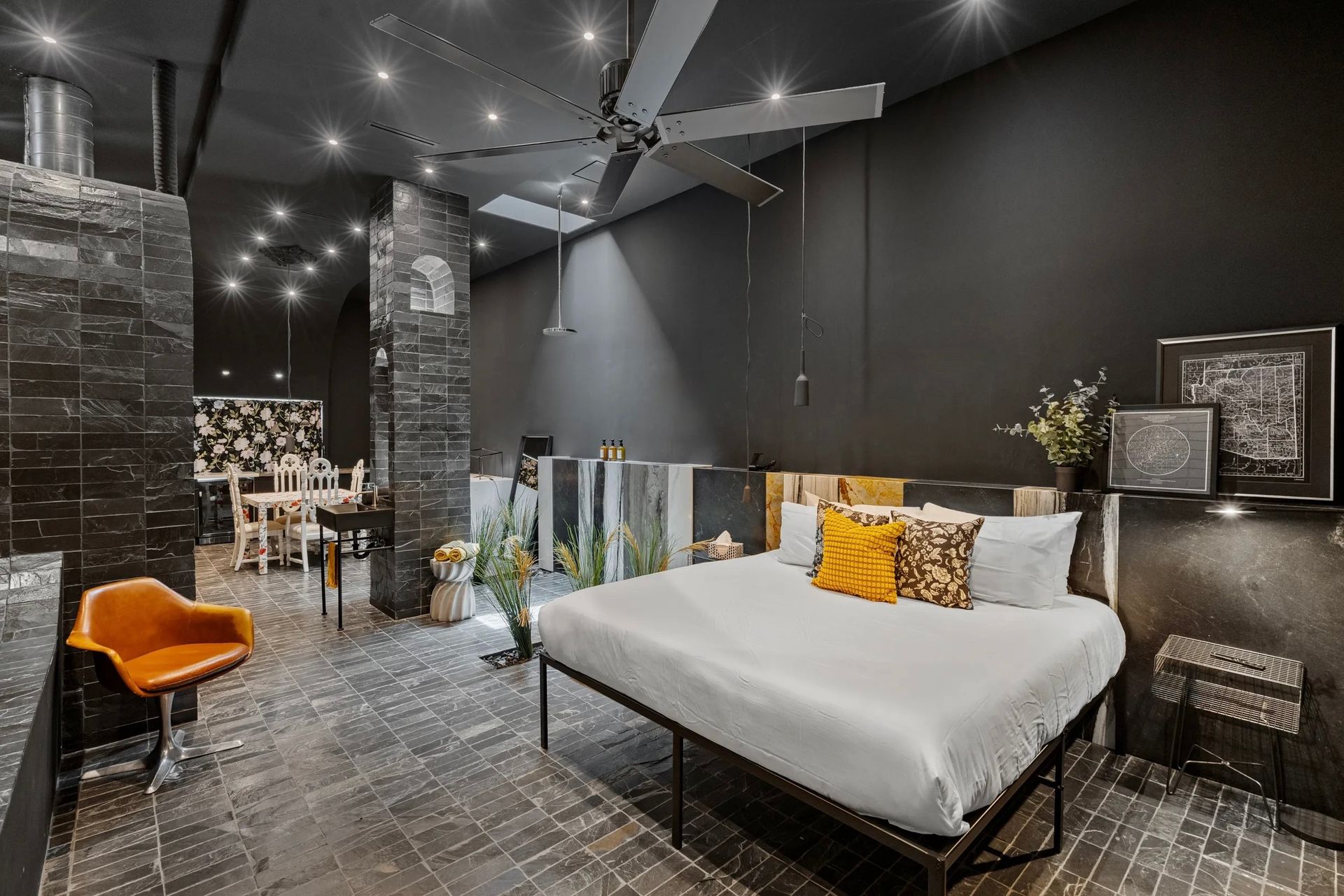 Dark, modern bedroom with a black wall, large bed, and an orange chair. There's a dining area visible in the background.