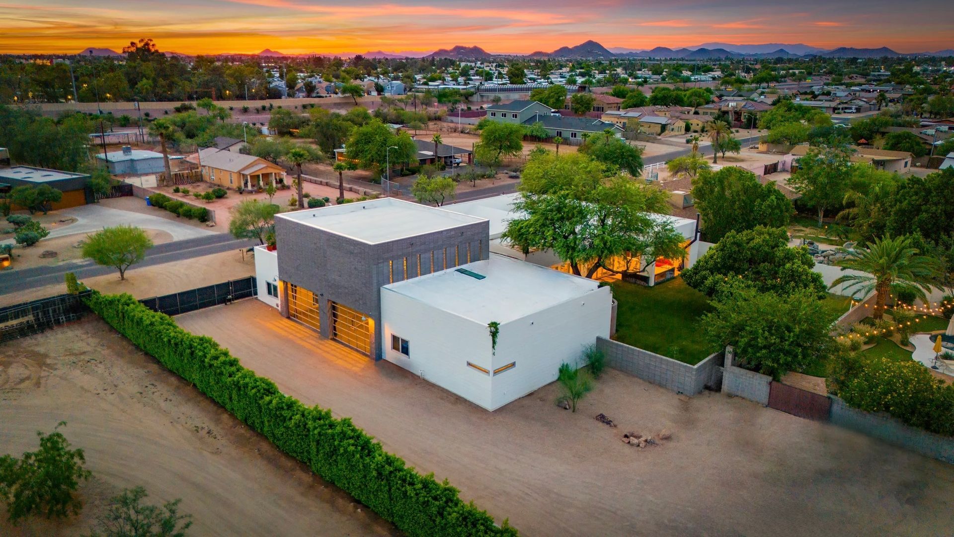 Aerial view of a modern white house with a flat roof, featuring a long green hedge and a sunset backdrop over a town.