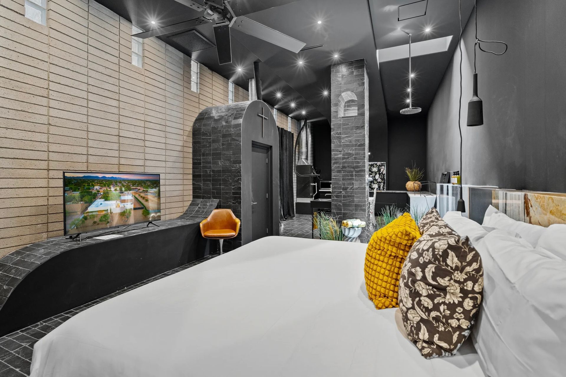 Modern bedroom with a large bed, black walls, and textured wood paneling. Includes a TV, seating, and a view of a shower.