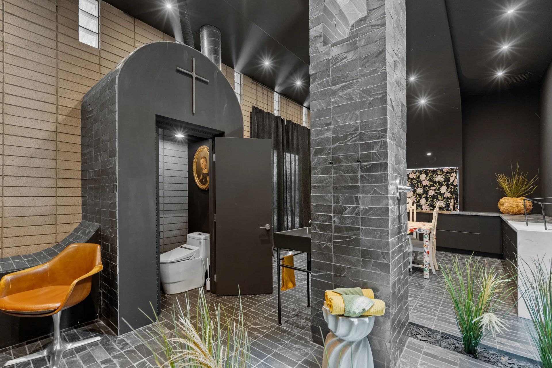A modern bathroom featuring a black arched toilet enclosure with a cross, a tan chair, and gray tiled walls.