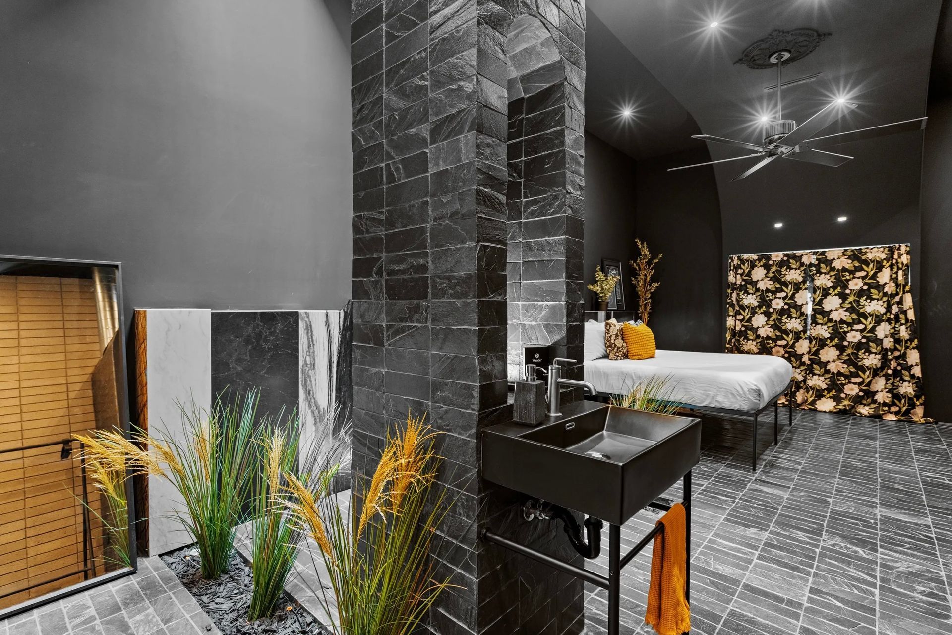 Dark spa interior with black tiled pillar, sink, and floral screen near a massage table, all in a moody atmosphere.