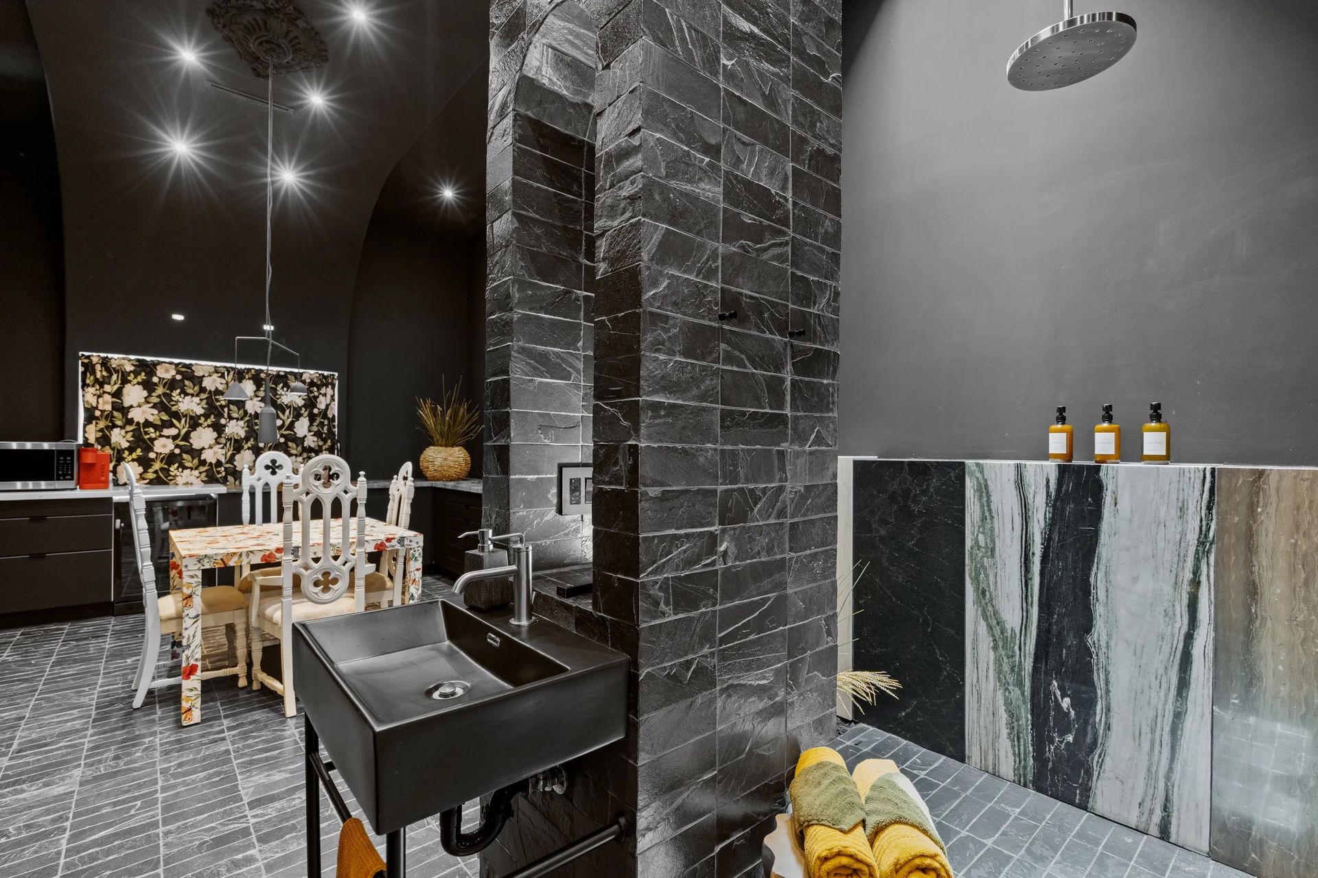 A modern bathroom with a dark color scheme. Features include black tile shower, black sink, and stone tiles on display.