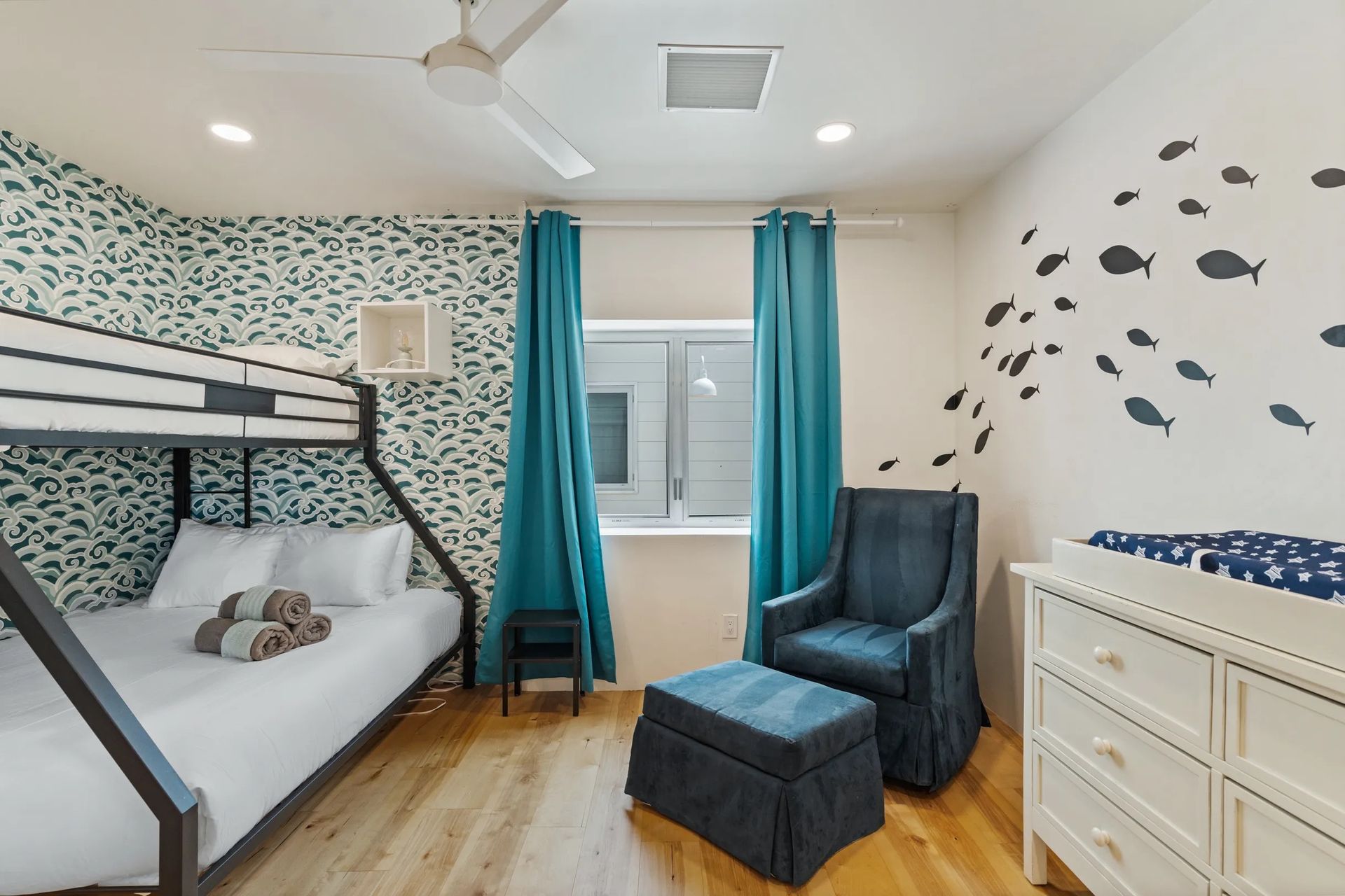 A bedroom with a bunk bed, teal curtains, a patterned accent wall, and a dresser with a changing pad.