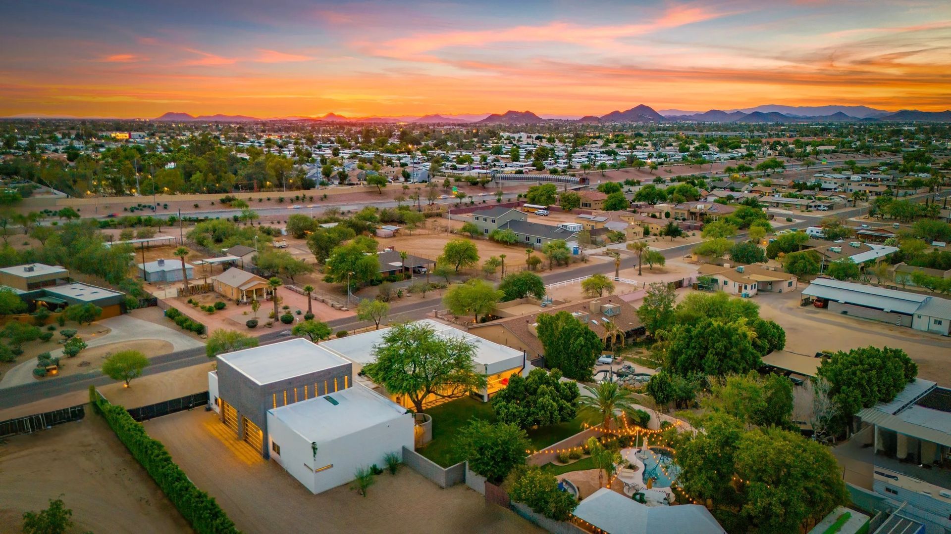 Aerial view of a suburban neighborhood at sunset, with modern white homes, green trees, and mountains in the distance against an orange sky.
