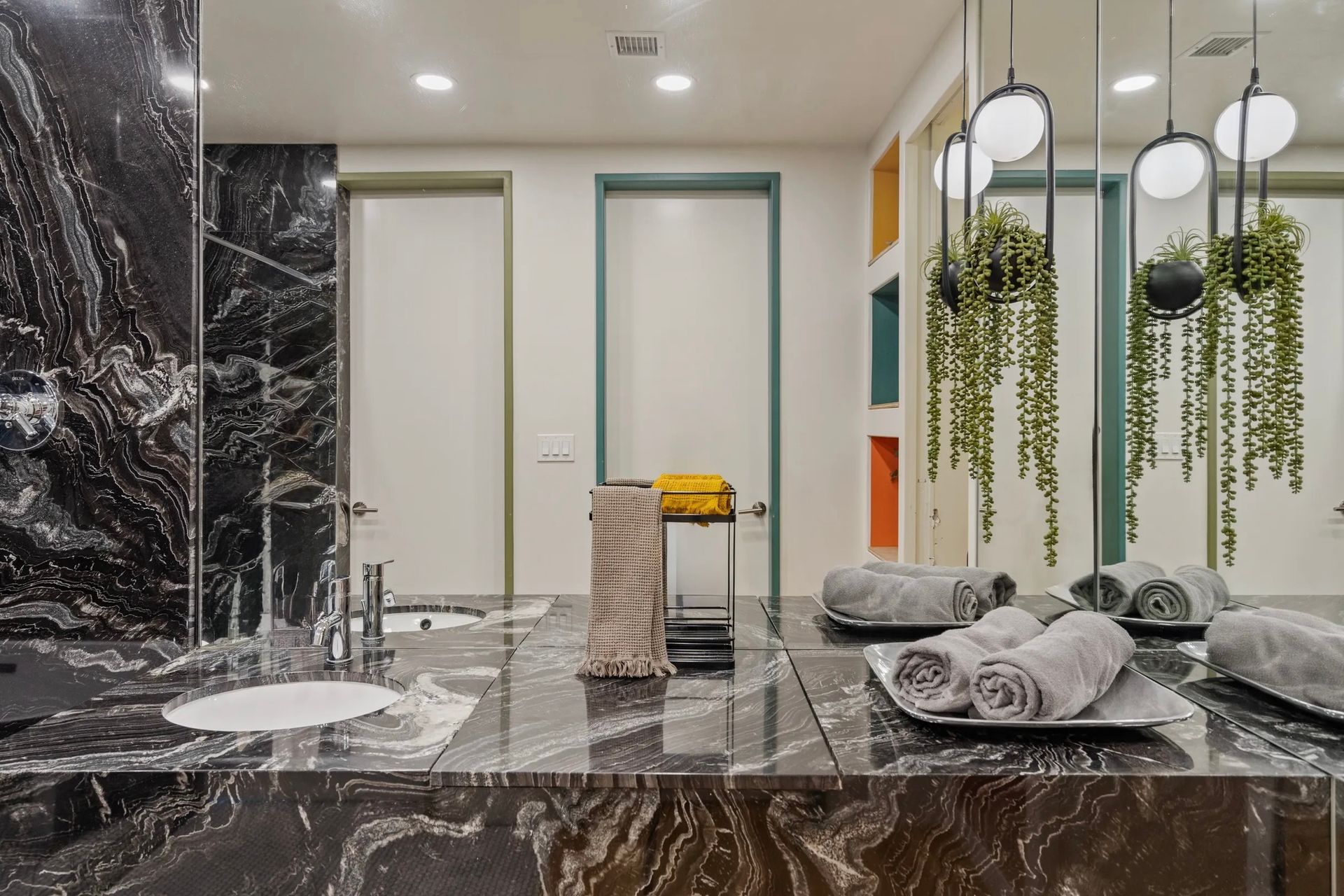 Bathroom with black marble countertop and backsplash, towels, hanging plants, and colorful doors in the background.