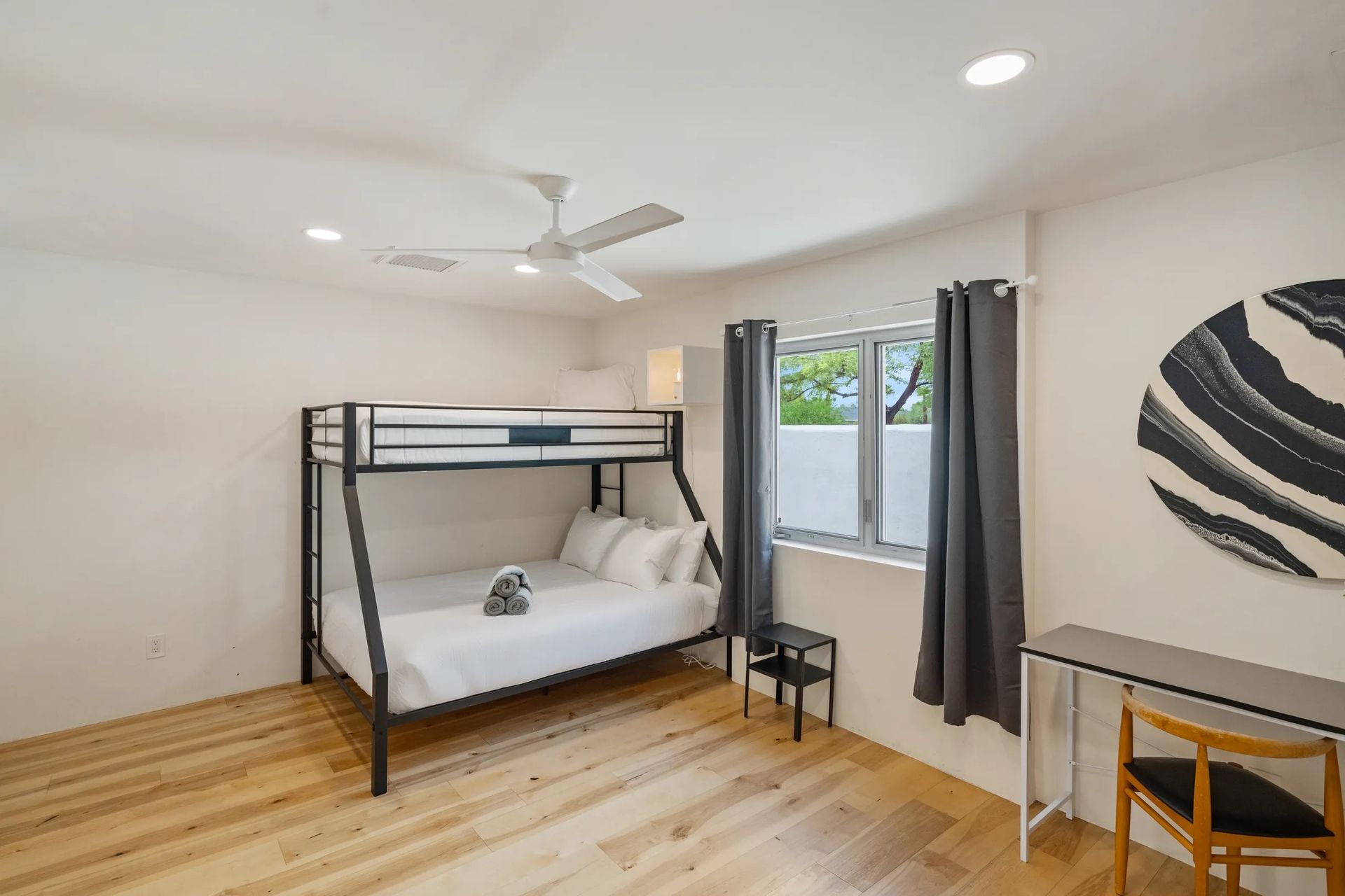 A bedroom with a black bunk bed, hardwood floors, and a window with gray curtains. There is a desk with a chair and a round wall decoration.