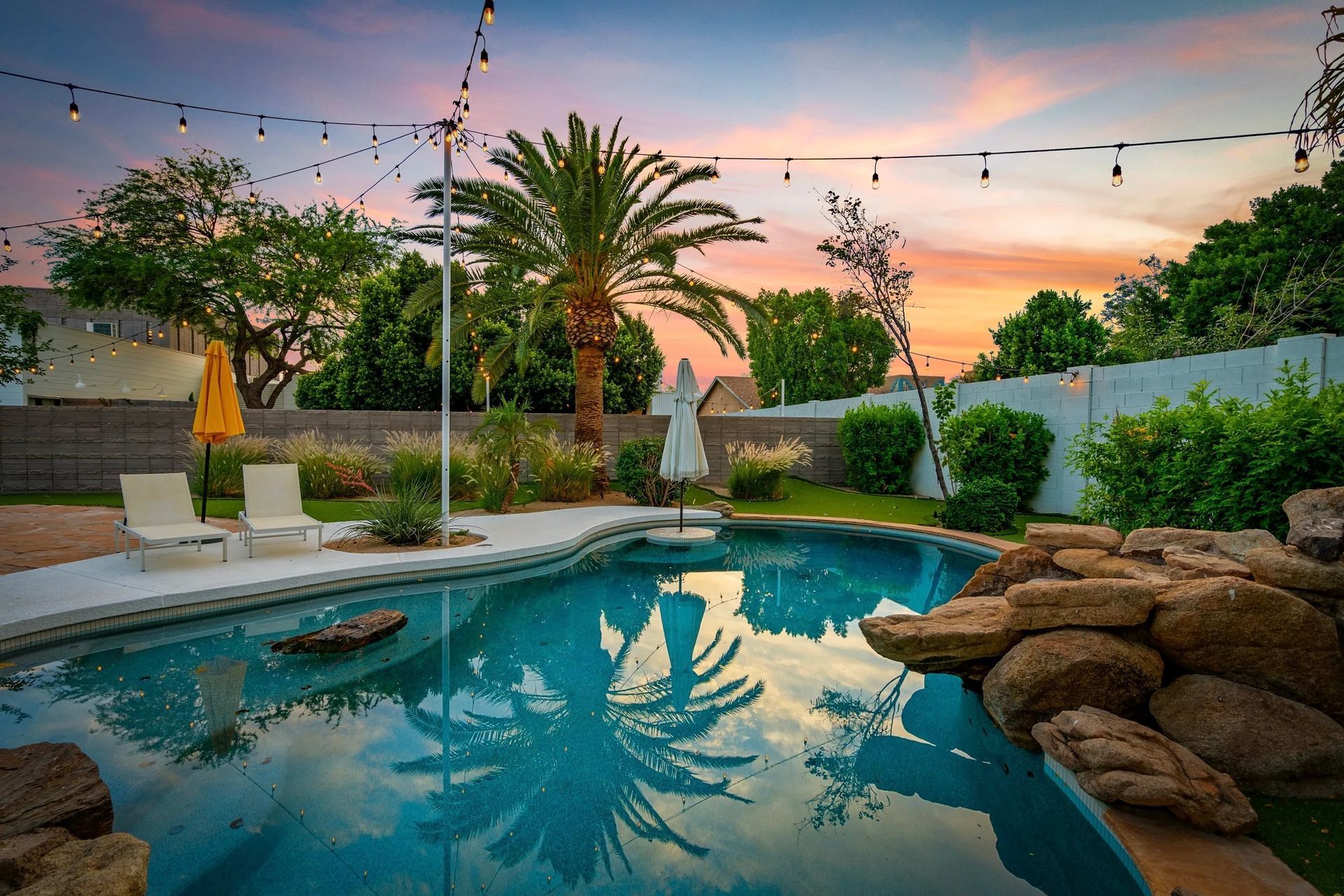 Backyard pool at sunset with string lights, palm tree, and lounge chairs. Sky is vibrant with pink, orange, and blue hues.