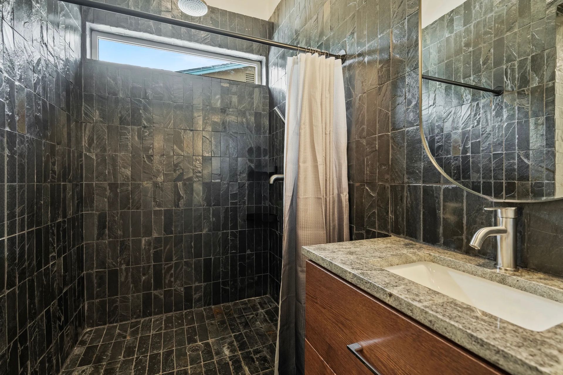 Modern bathroom with dark tile walls, a walk-in shower, a wooden vanity with a granite countertop, and a round mirror.