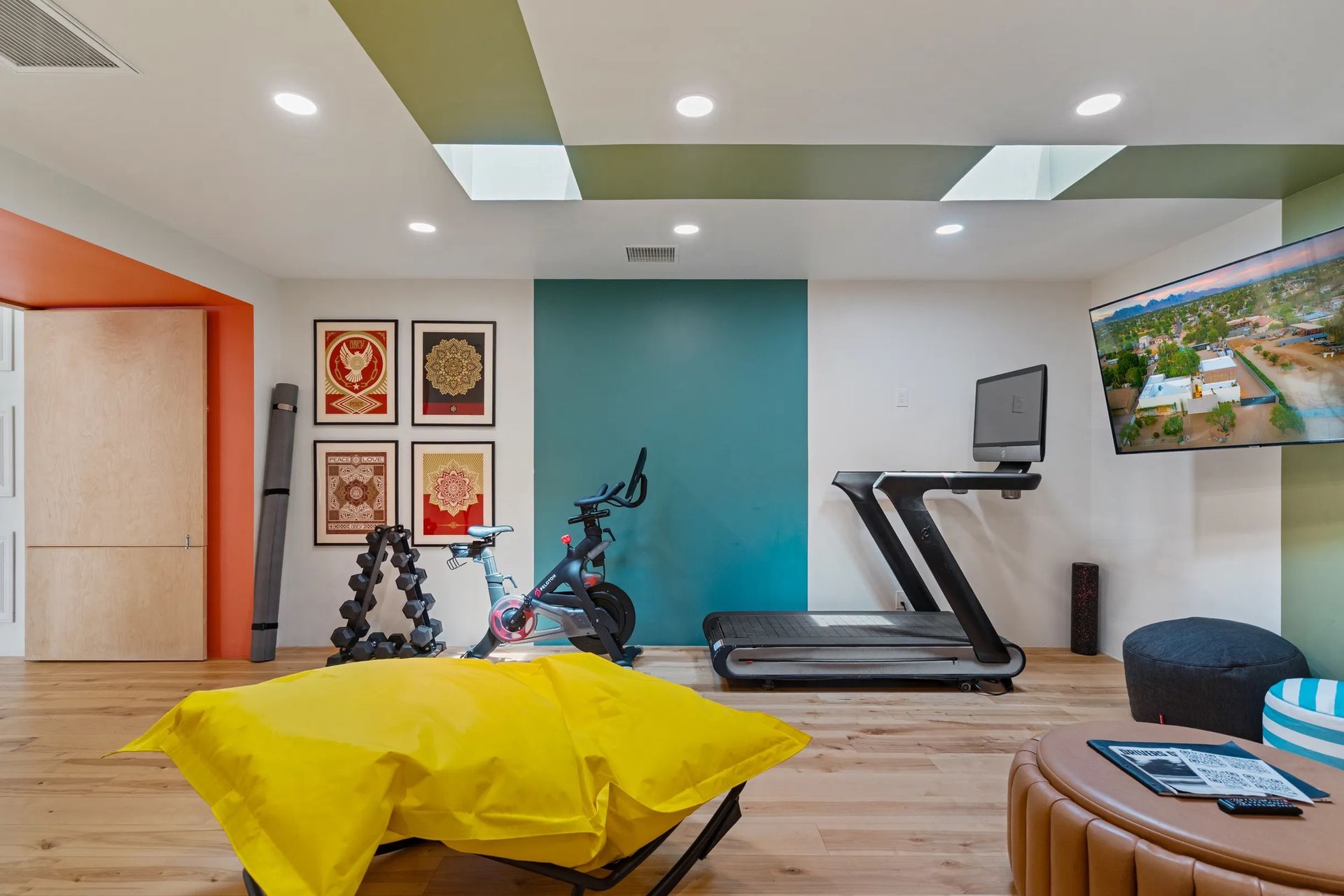 A home gym with exercise equipment, a mounted TV, and colorful accents on the walls and ceiling. Wooden floor and yellow beanbag.