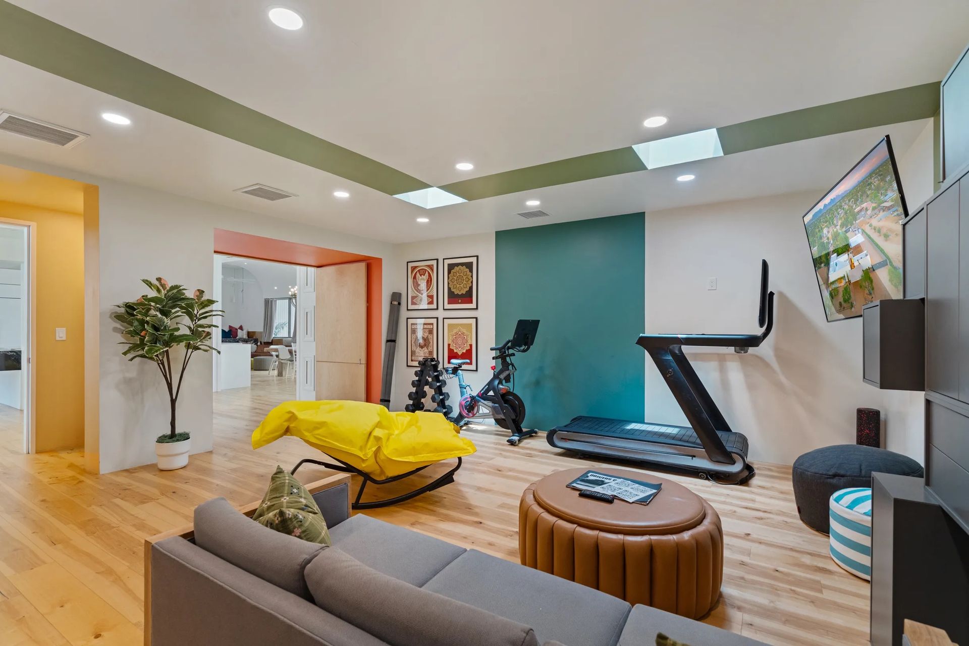 A modern home gym with a treadmill, exercise bike, seating, and a TV. The room features light wood flooring and a teal accent wall.