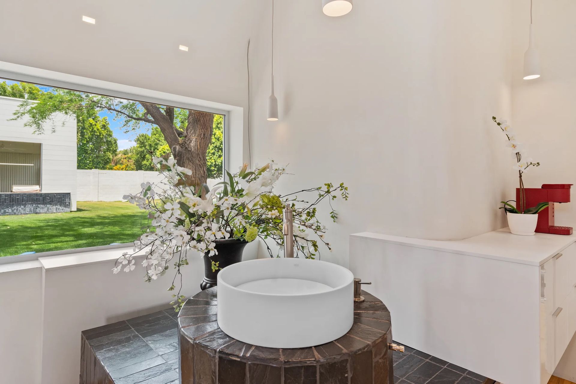 A modern bathroom with a white sink on a dark pedestal. A window overlooks a green lawn and trees.