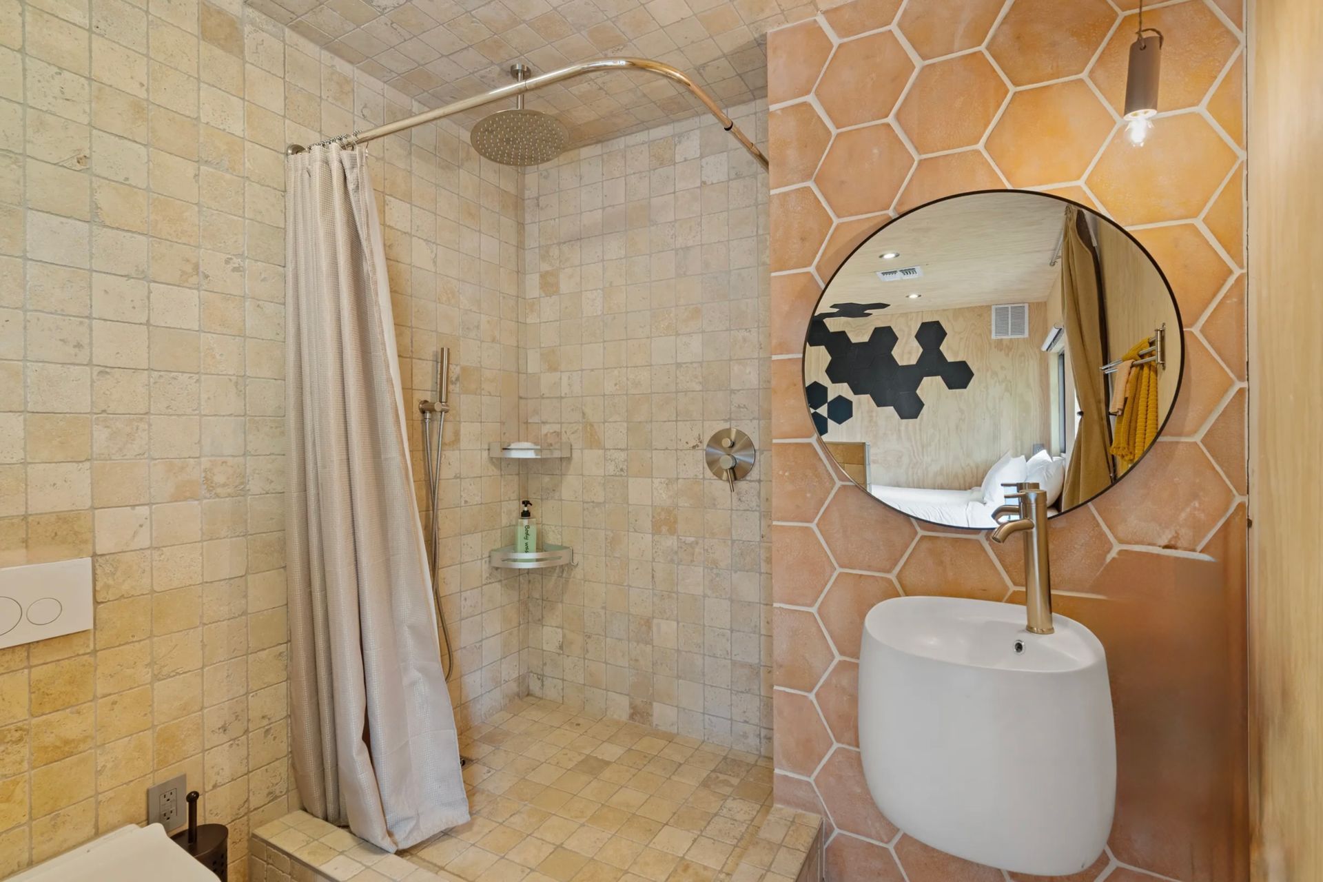 Small bathroom with peach-colored hexagonal tiles. A shower with a cream-colored curtain, a round mirror, and a white sink are visible.