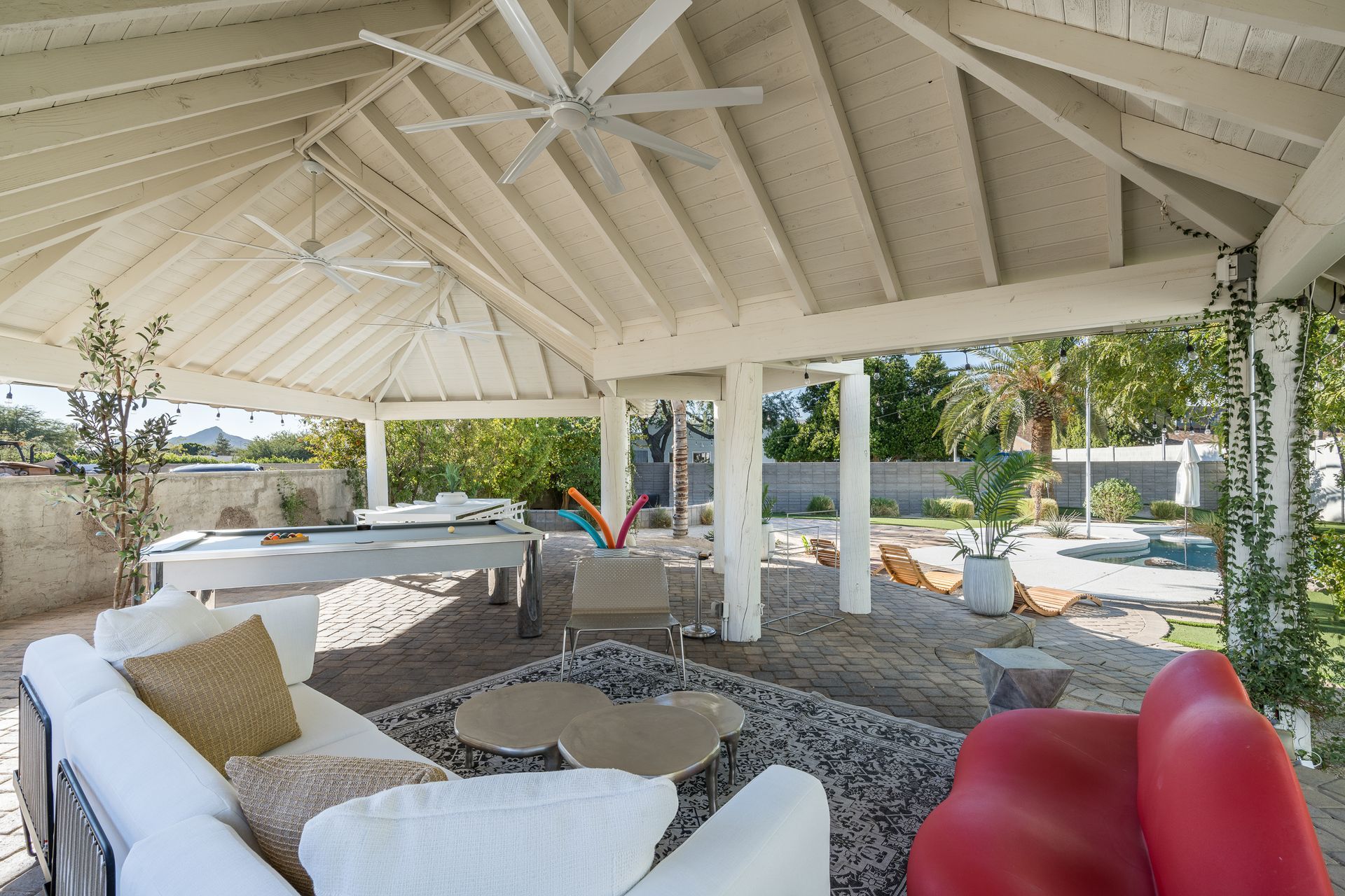 Covered patio with white beams and furniture, including a white couch with gold pillows and a red sofa. Overlooking a yard.