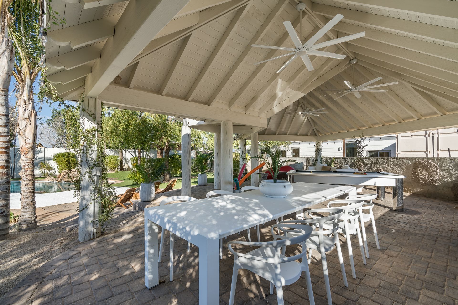 Outdoor dining area with a long white table and chairs under a white-painted wooden roof with fans.