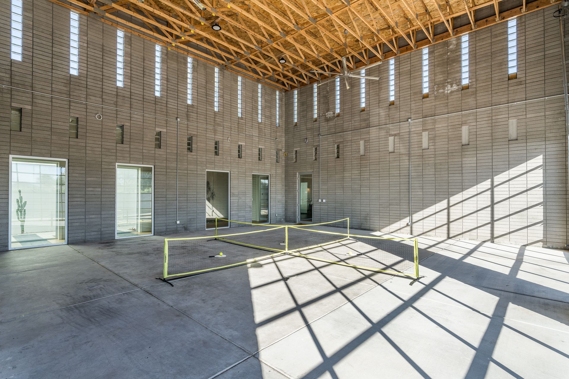 Large, empty industrial space with concrete walls, rows of vertical windows, and a wooden beam ceiling. Sunlight casts shadows across the floor.