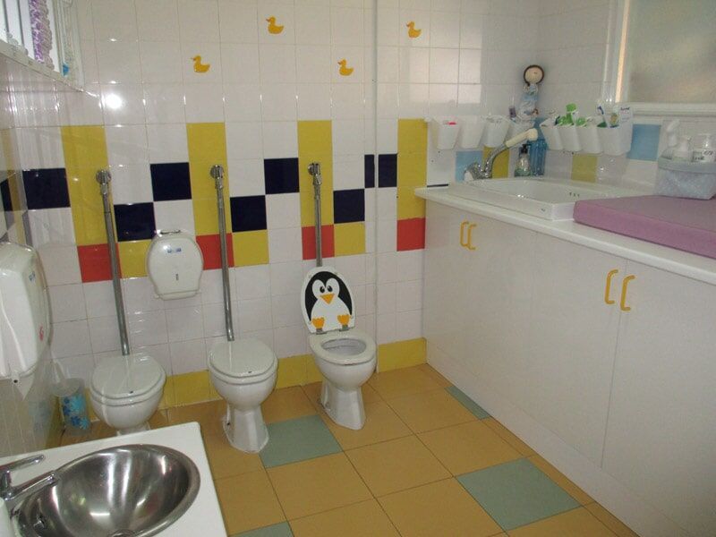 Banheiro infantil com três vasos sanitários pequenos, uma pia, trocador e azulejos com design lúdico.