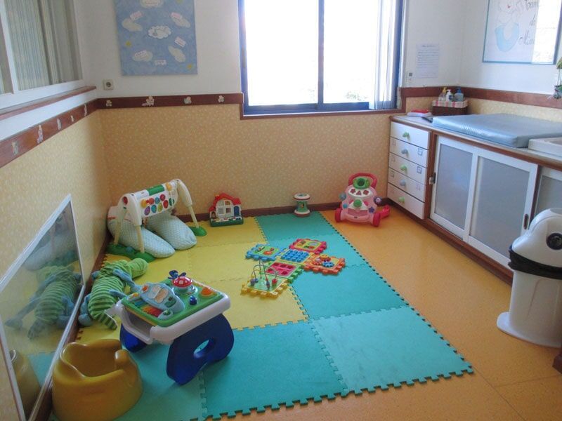 Área de recreação infantil com brinquedos em tapetes coloridos, ao lado de um trocador de fraldas e uma janela.