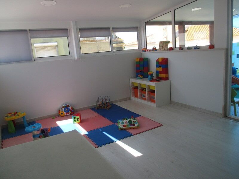 Sala de jogos com brinquedos em tapetes perto das janelas; paredes brancas, piso de madeira.