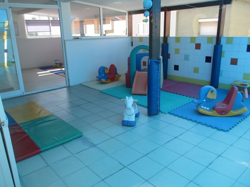 Sala de jogos com tapetes coloridos, estruturas acolchoadas e brinquedos de montar sobre piso de azulejo.