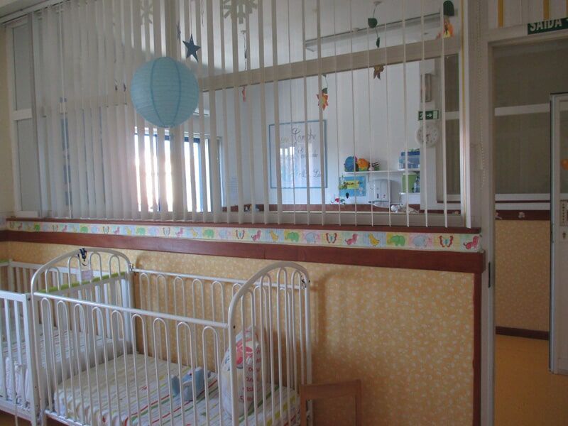 Quarto de bebê com berços, persianas e faixa decorativa. Uma esfera azul está pendurada no teto.